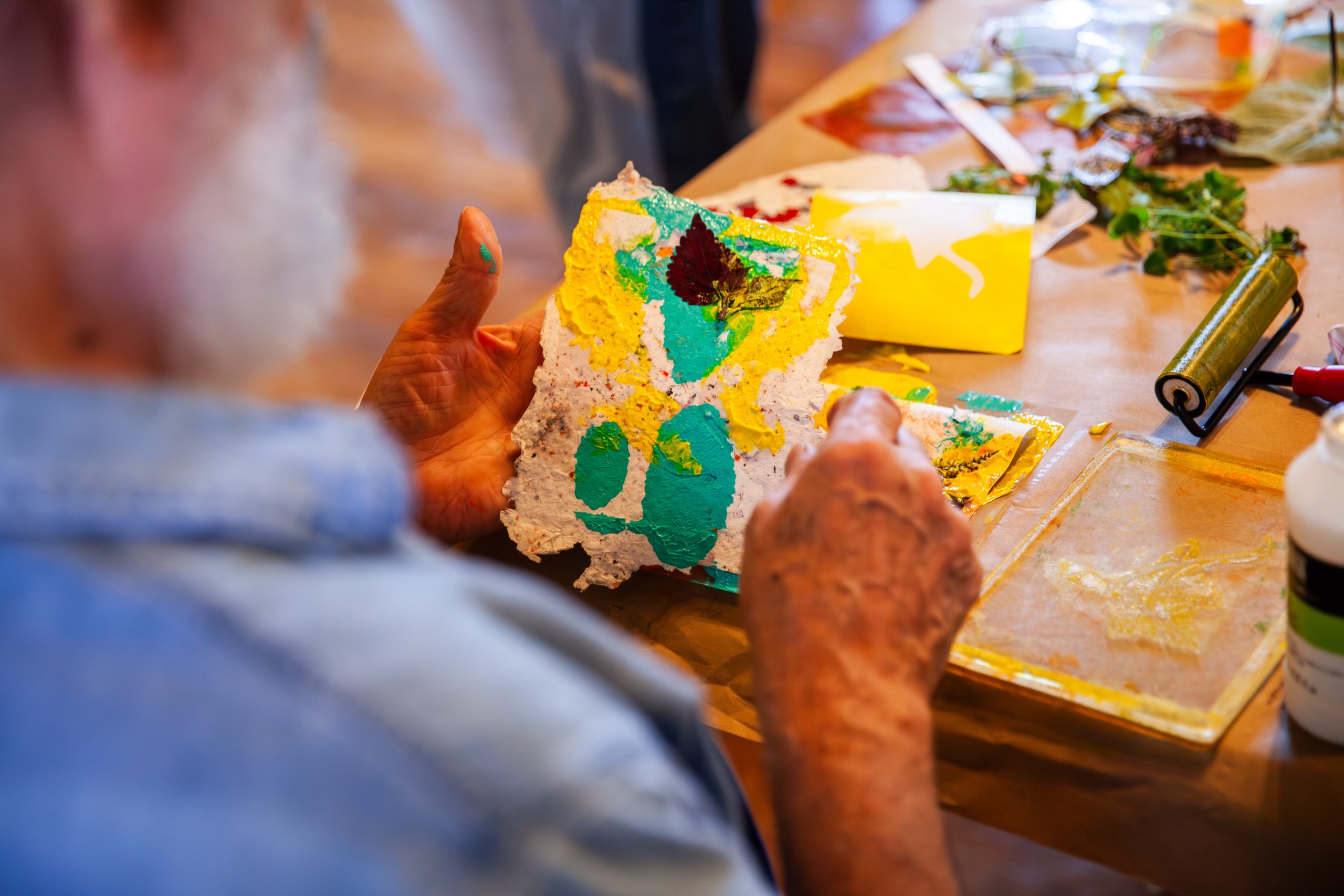 A person with a white beard holds a textured piece of paper decorated with bright green and yellow paint, working at a cluttered art table with paints, rollers, and leaves.
