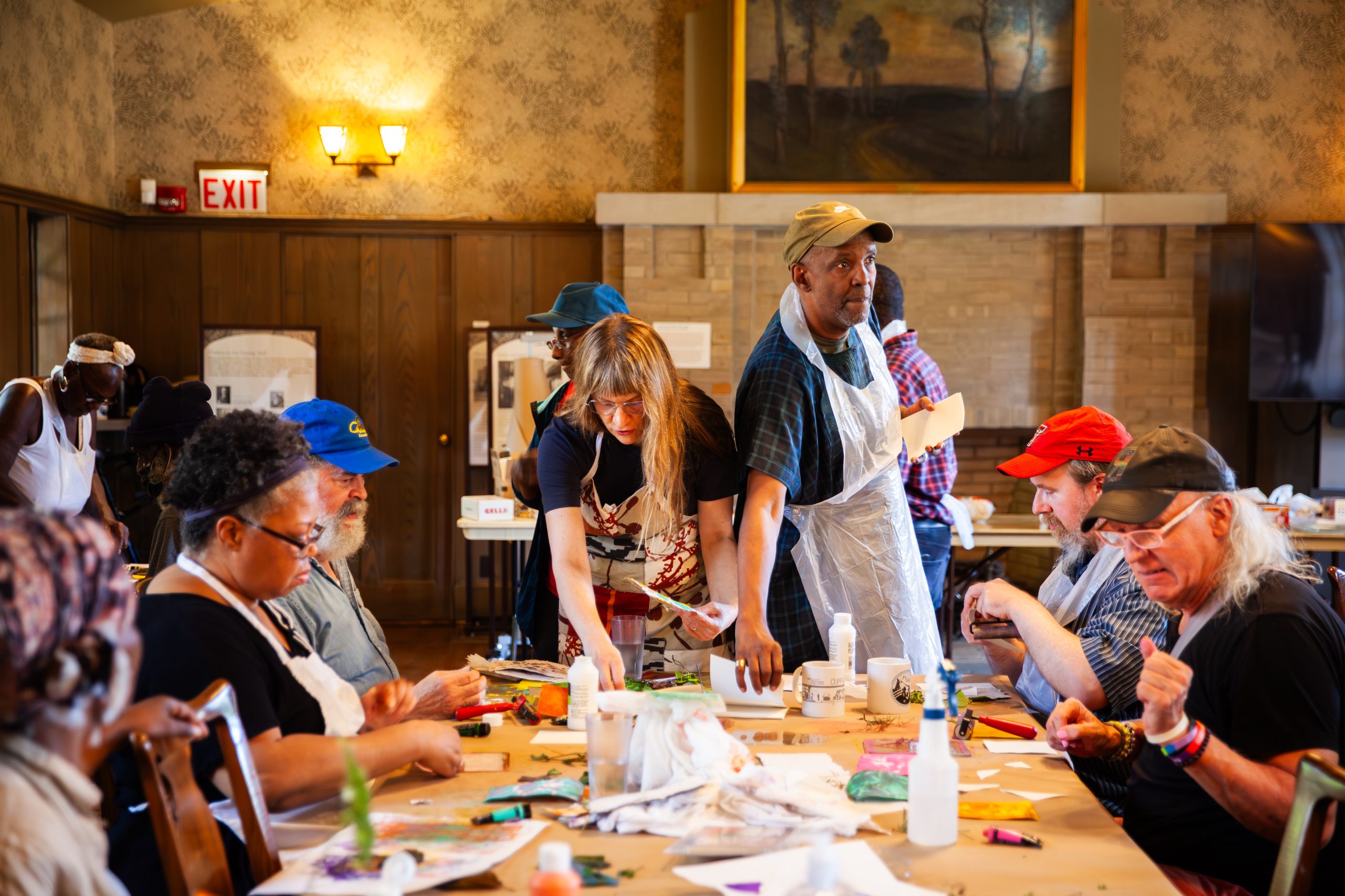 A group of adults sits around a table engaged in a lively arts and crafts activity, with paint, brushes, and paper scattered on the table in a warmly lit room. Some people are standing and assisting others.