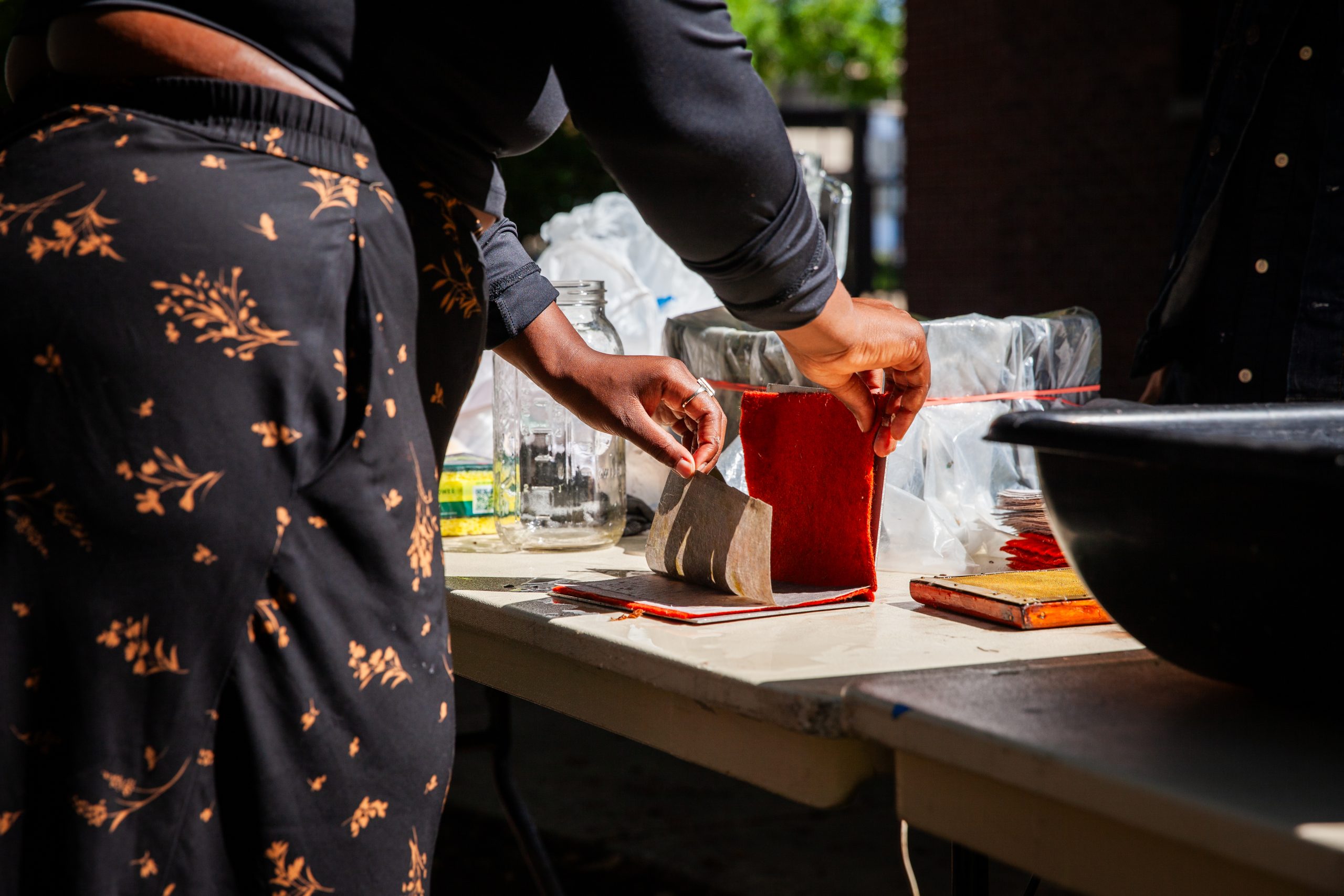 A person stands at a table outdoors, holding a piece of brown paper and dipping it into a rectangular container with red edges, possibly involved in a creative or craft activity.