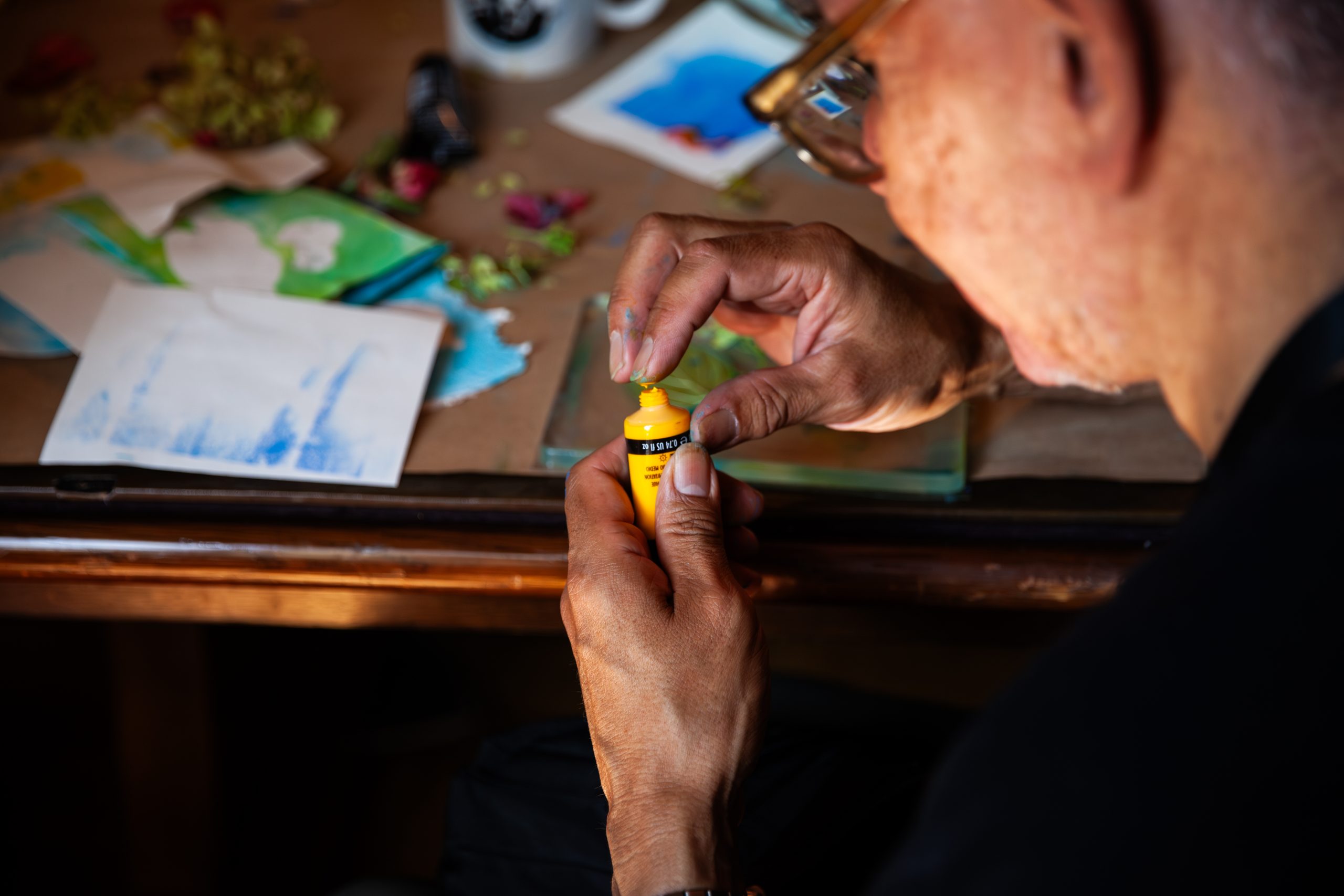 A person wearing glasses holds a small yellow tube of watercolor paint above a table scattered with art supplies, papers, and colorful artworks in progress.