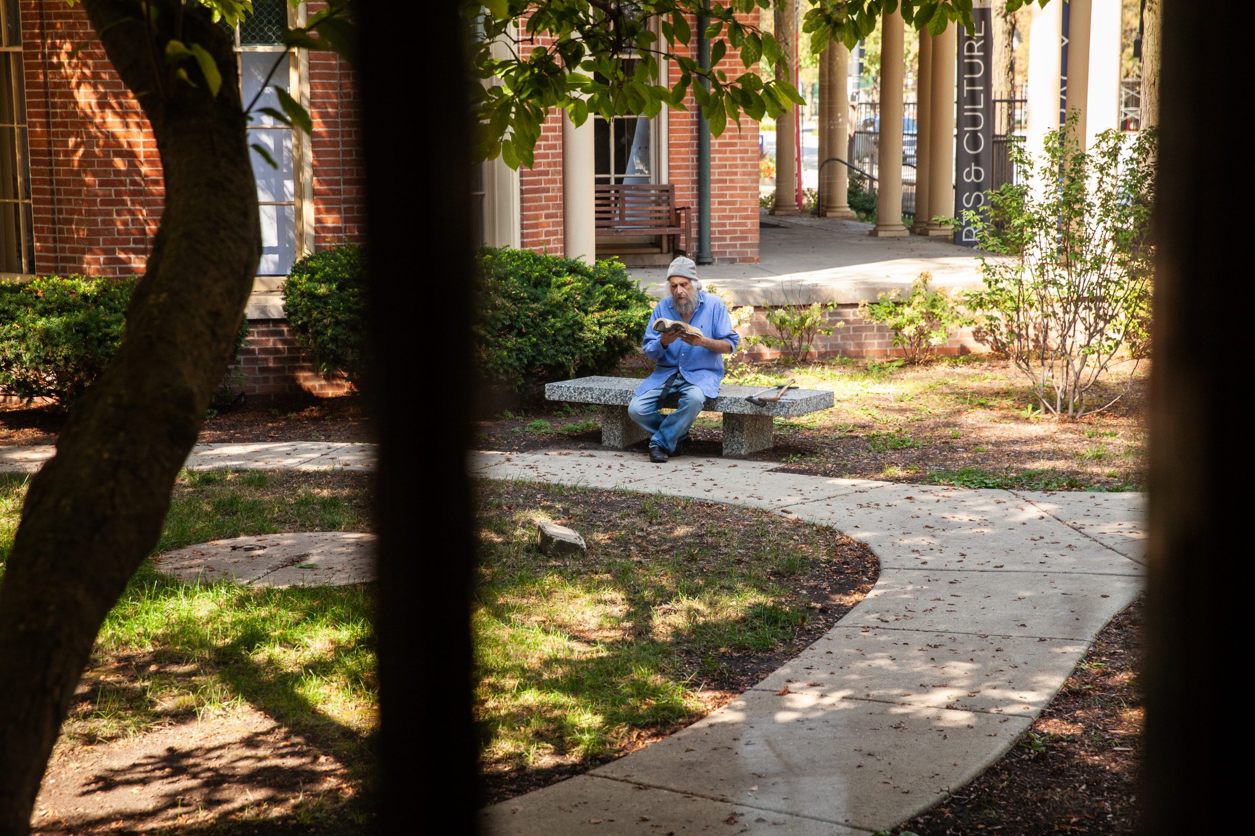 A man in a blue shirt and cap sits alone on a stone bench in a courtyard, reading a book. Curved walkways and trees surround him, and sunlight filters through the foliage.
