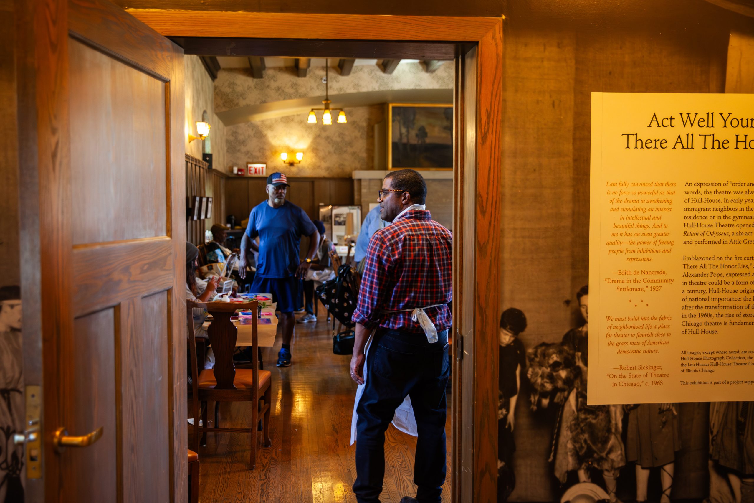 Two men stand in the doorway of a wood-paneled room filled with people seated at tables. Warm lighting, framed art, and a large text display are visible on the wall near the entrance.