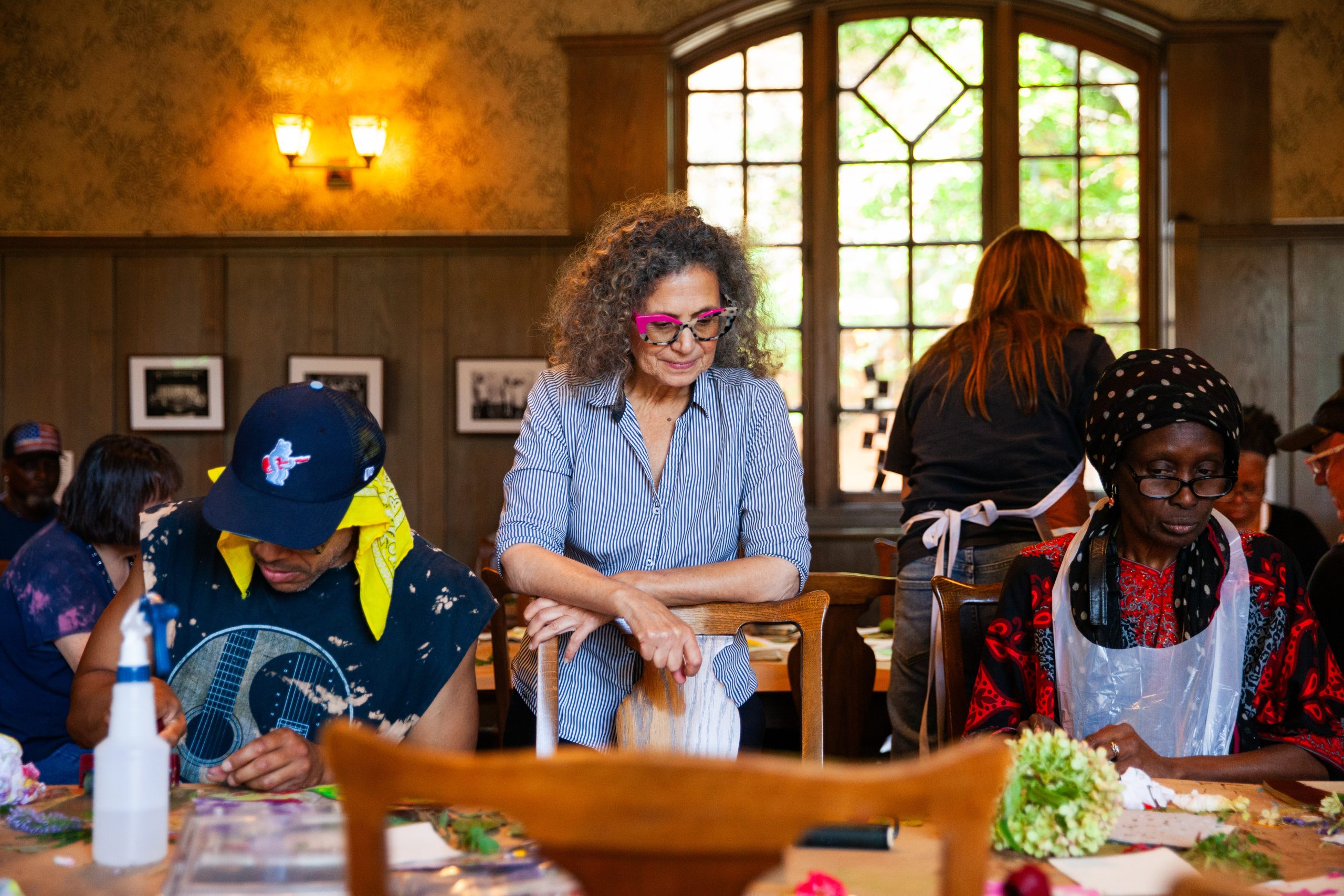 A woman with curly hair and pink glasses stands behind two people sitting at a table doing crafts. The room has wooden walls, framed photos, and a large window letting in natural light.