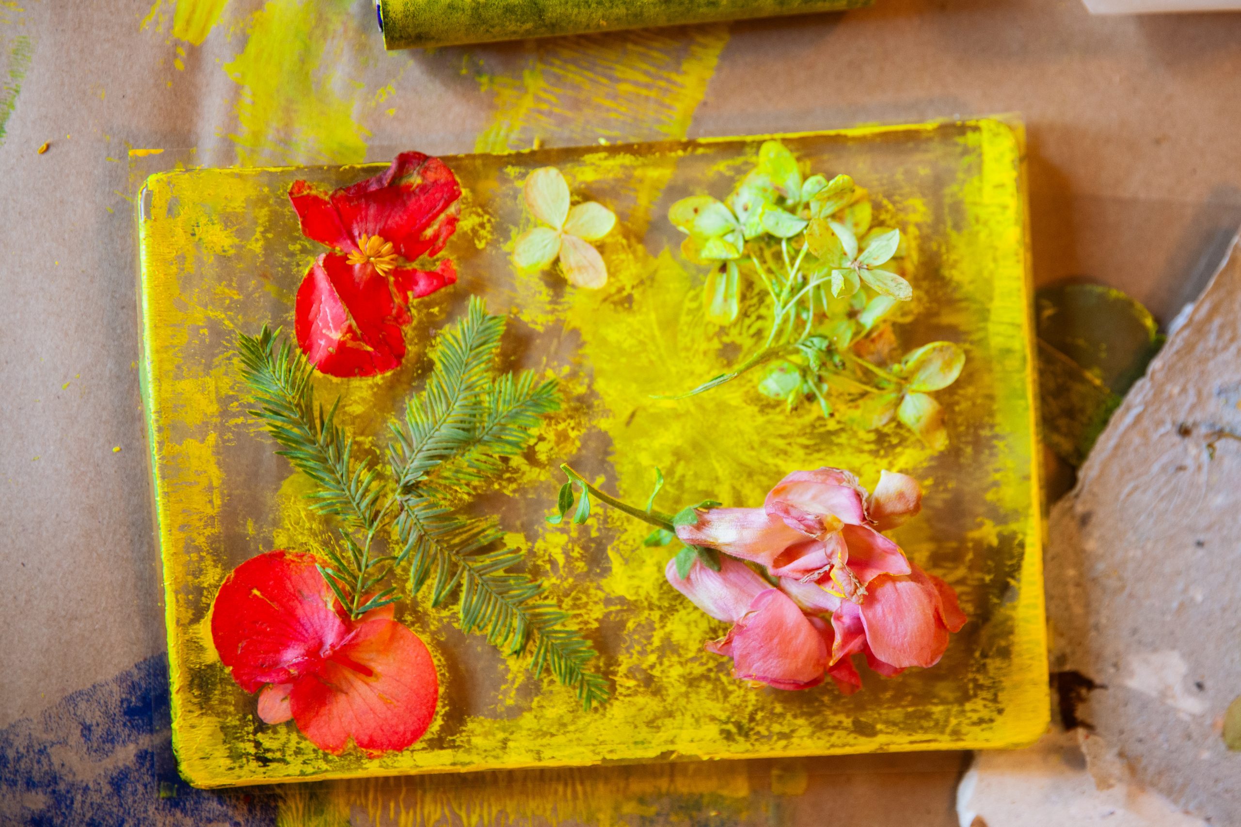 A yellow-painted surface with red and pink flowers, green leaves, and a spruce twig arranged on top, set on a brown paper-covered table with scattered paint marks.