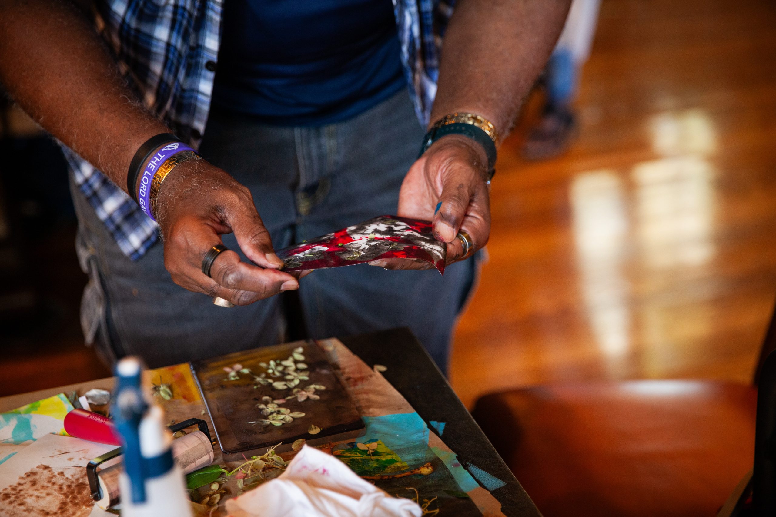 A person holds a small, abstractly painted canvas or paper above a table with art supplies, paint, and tissue, suggesting they are engaged in creating art. Only their hands and torso are visible.