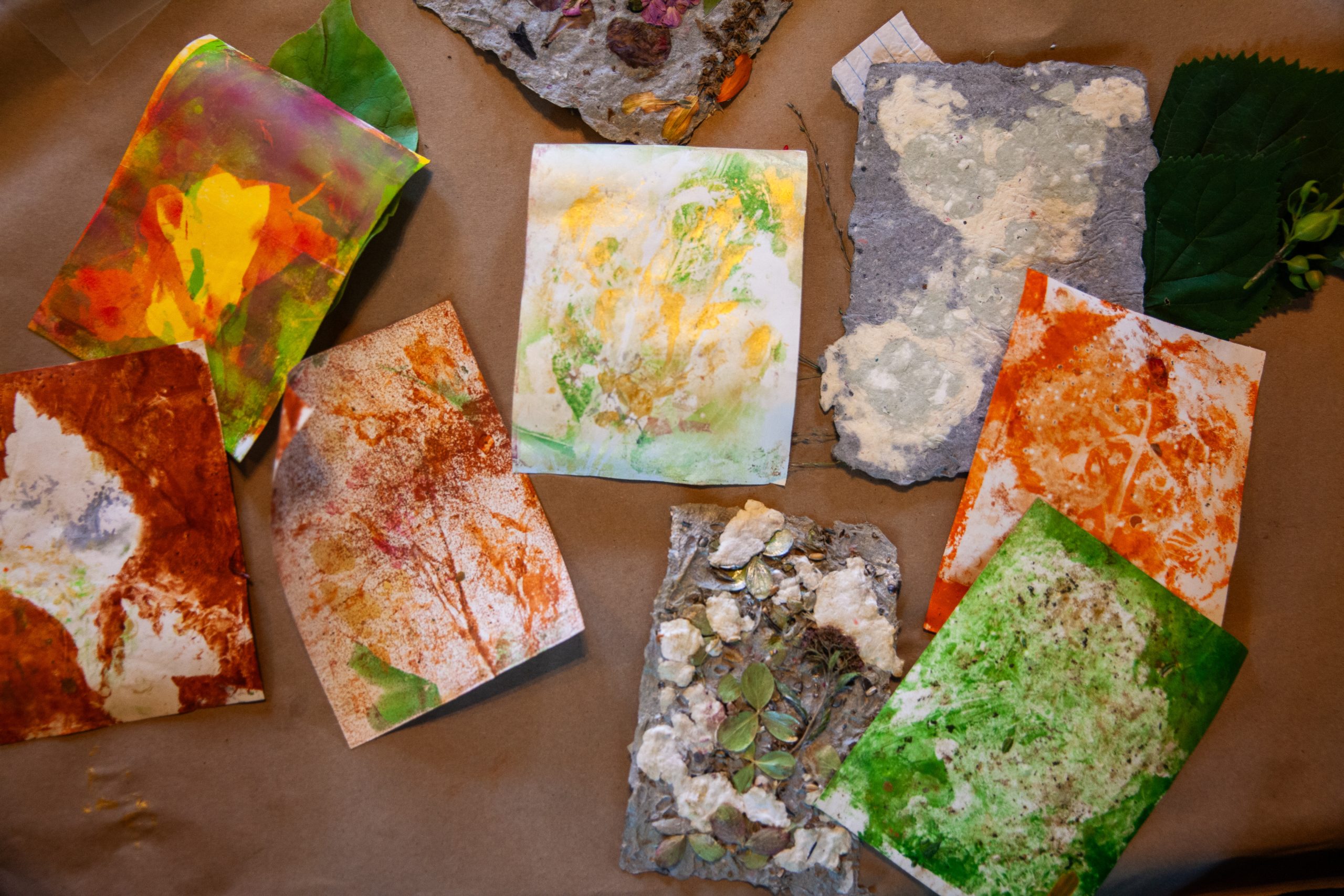 Several handmade papers and colorful painted cards with abstract designs are spread out on a brown surface. Some papers have pressed flowers or leaves, while others display vivid red, green, and yellow paint patterns.