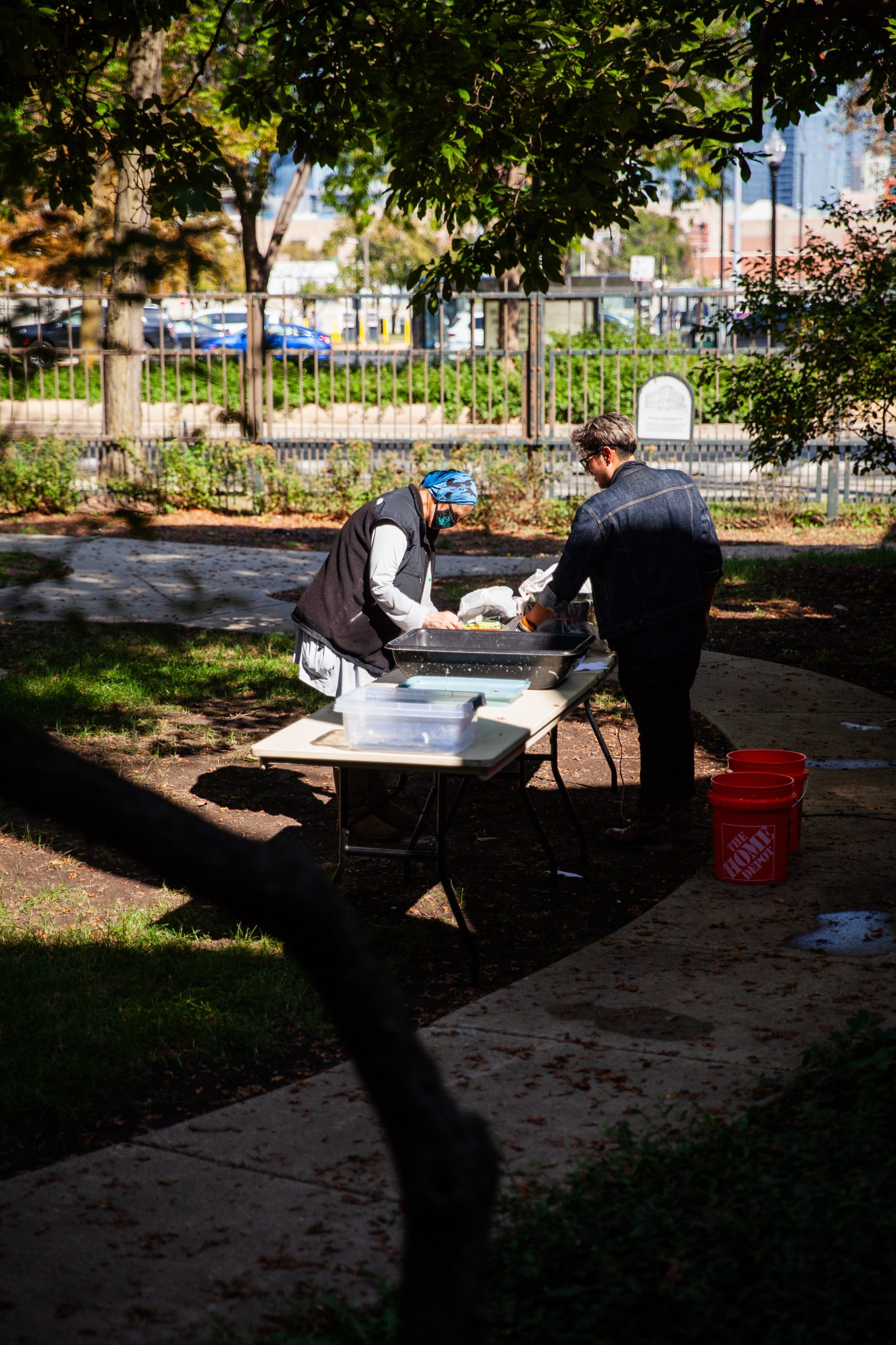 Two people stand at an outdoor table preparing food or supplies under trees in a park. Large plastic bins and orange buckets are on or near the table. Sunlight and shadows fall across the scene. A fence and parked cars are in the background.