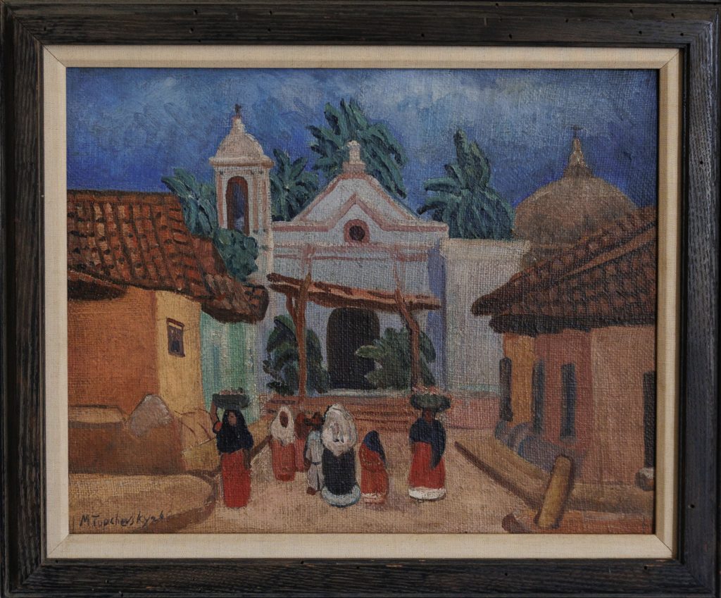 A framed painting depicts a village scene with women in colorful skirts and shawls walking toward a white church, surrounded by rustic houses with red-tiled roofs and palm trees in the background under a blue sky.