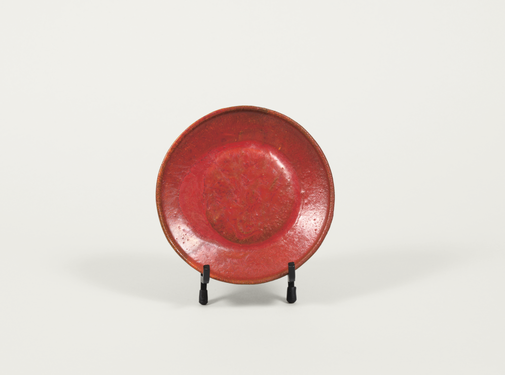 A round, shallow red plate with a slightly raised rim, displayed upright on a black stand against a plain white background. The plate has a smooth, glossy surface with subtle texture variations.