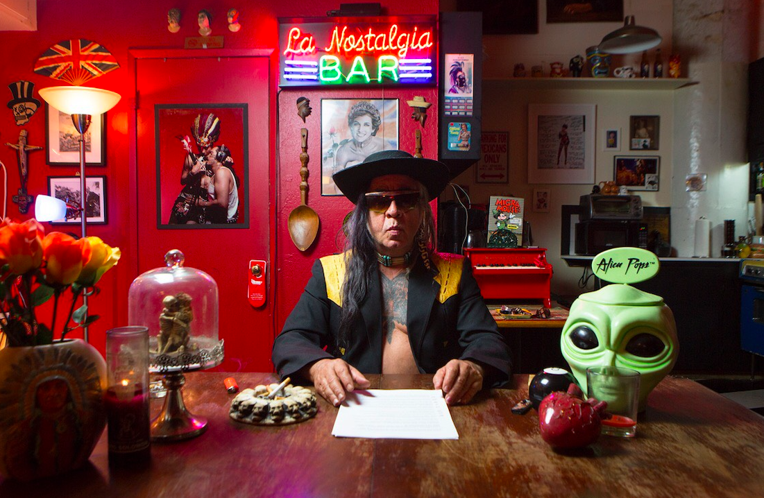 A man wearing sunglasses and a black hat sits at a cluttered table in a colorful room with eclectic decor, including flowers, candles, a green alien head, and a neon sign reading La Nostalgia Bar above him.