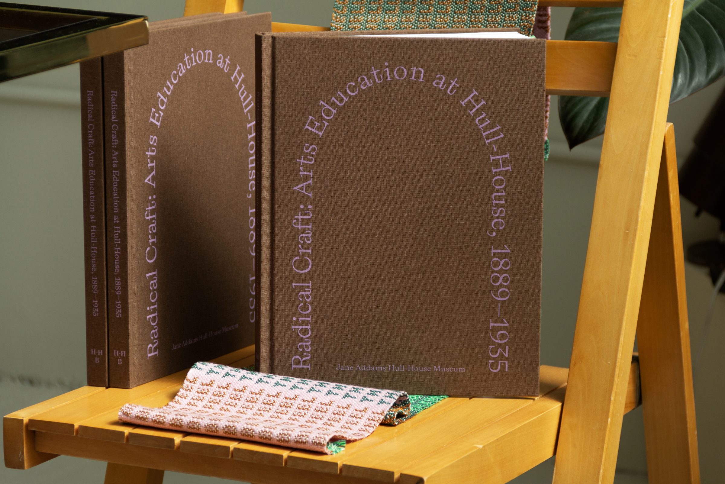 A brown book titled “Radical Craft: Arts Education at Hull House, 1889–1935” is displayed on a wooden chair, with another similar book beside it and a folded patterned fabric resting on the seat.
