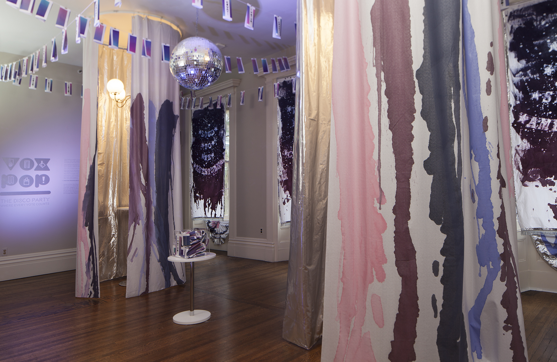 Art installation in a room with flowing fabric panels painted in blue, pink, and purple tones, a mirrored disco ball, flag banners, a vintage lamp, and a small round table with books; sunlight streams through painted windows.
