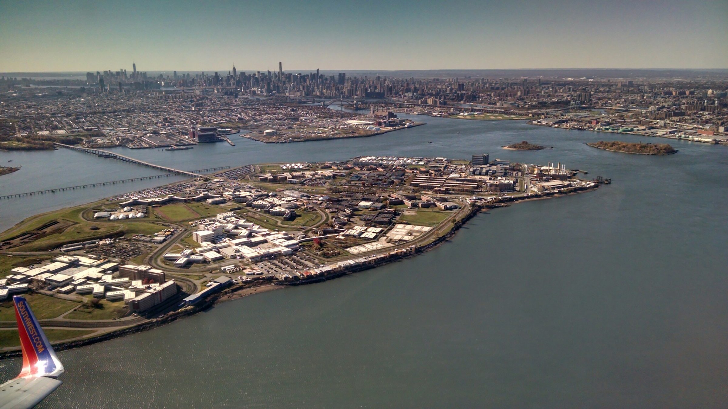 Aerial view of Rikers Island in New York City, surrounded by water with the skyline of Manhattan visible in the background under a clear sky.