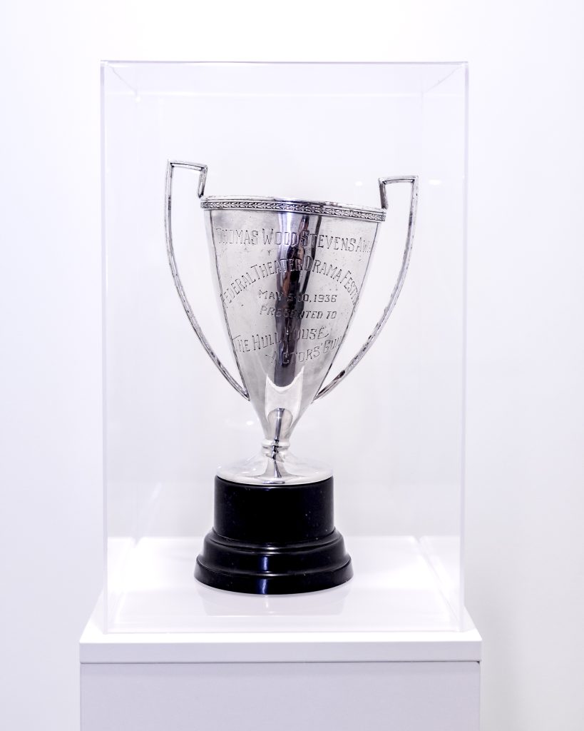 A silver trophy with two handles and engraved text is displayed in a clear glass case on a white pedestal against a plain white background.