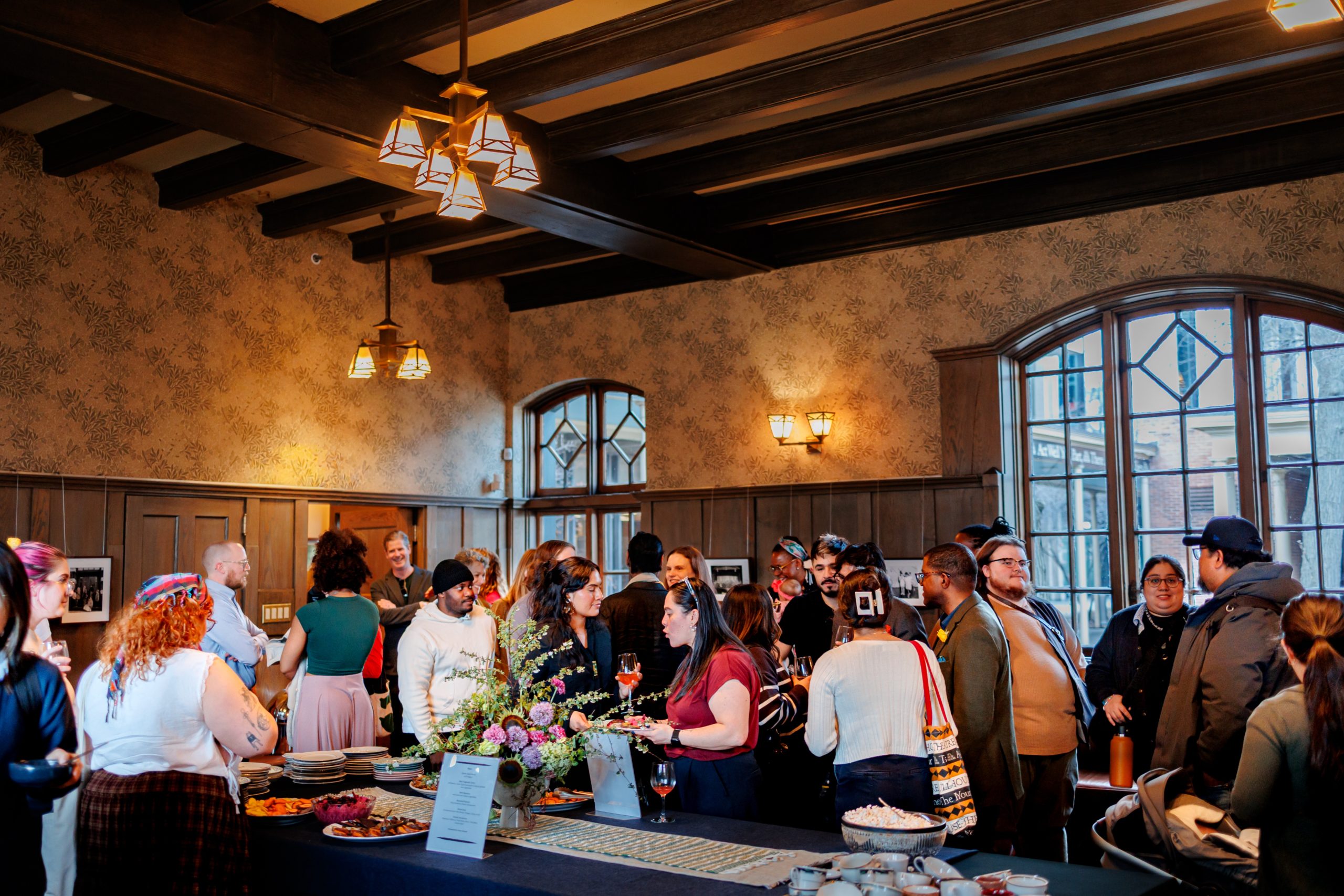 A group of people gather in a warmly lit room with wooden beams and large windows, socializing around a long table filled with food, flowers, and drinks. The atmosphere is lively and festive.