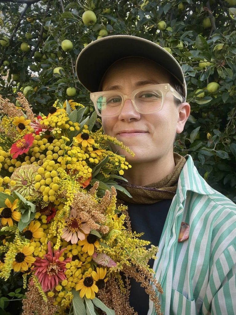 A person wearing glasses, a cap, and a green striped shirt holds a colorful bouquet of yellow and pink flowers, standing in front of a leafy apple tree with green apples.