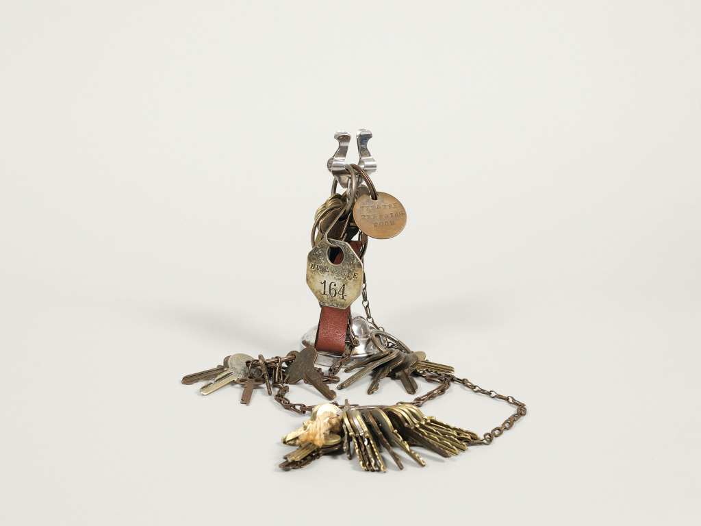 A vintage jumble of assorted keys, brass tags, a metal chain with a hand-shaped pendant, and a leather key fob marked 164, all arranged against a plain white background.