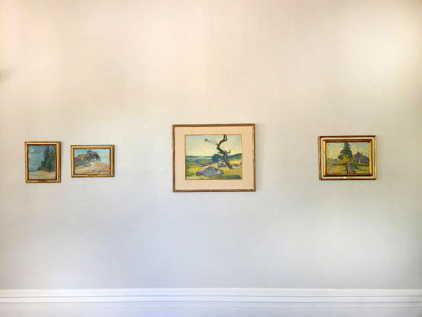 Four framed landscape paintings hang on a plain, light-colored wall. The central painting features a stag; the others depict trees and natural scenes. A white baseboard runs along the bottom of the wall.