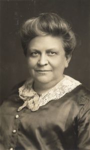 A black-and-white portrait of an older woman with short, wavy hair, wearing a satin dress with buttons and a lace collar, and a small scarf tied at her neck. She is facing the camera and smiling slightly.