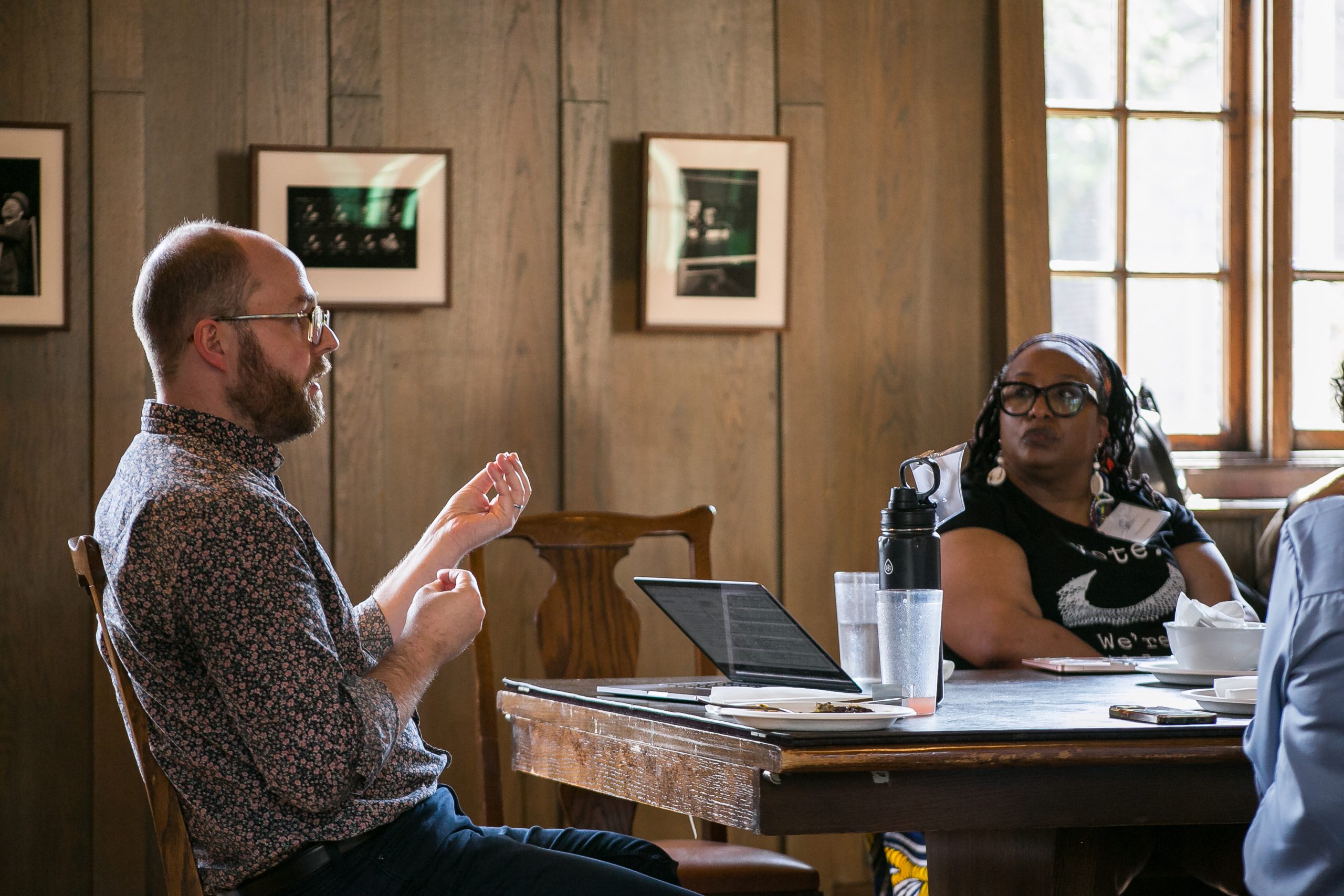A man speaks while gesturing with his hands at a wooden table with a laptop, as a woman listens attentively. Framed photos hang on the wall behind them. Sunlight streams through a window.