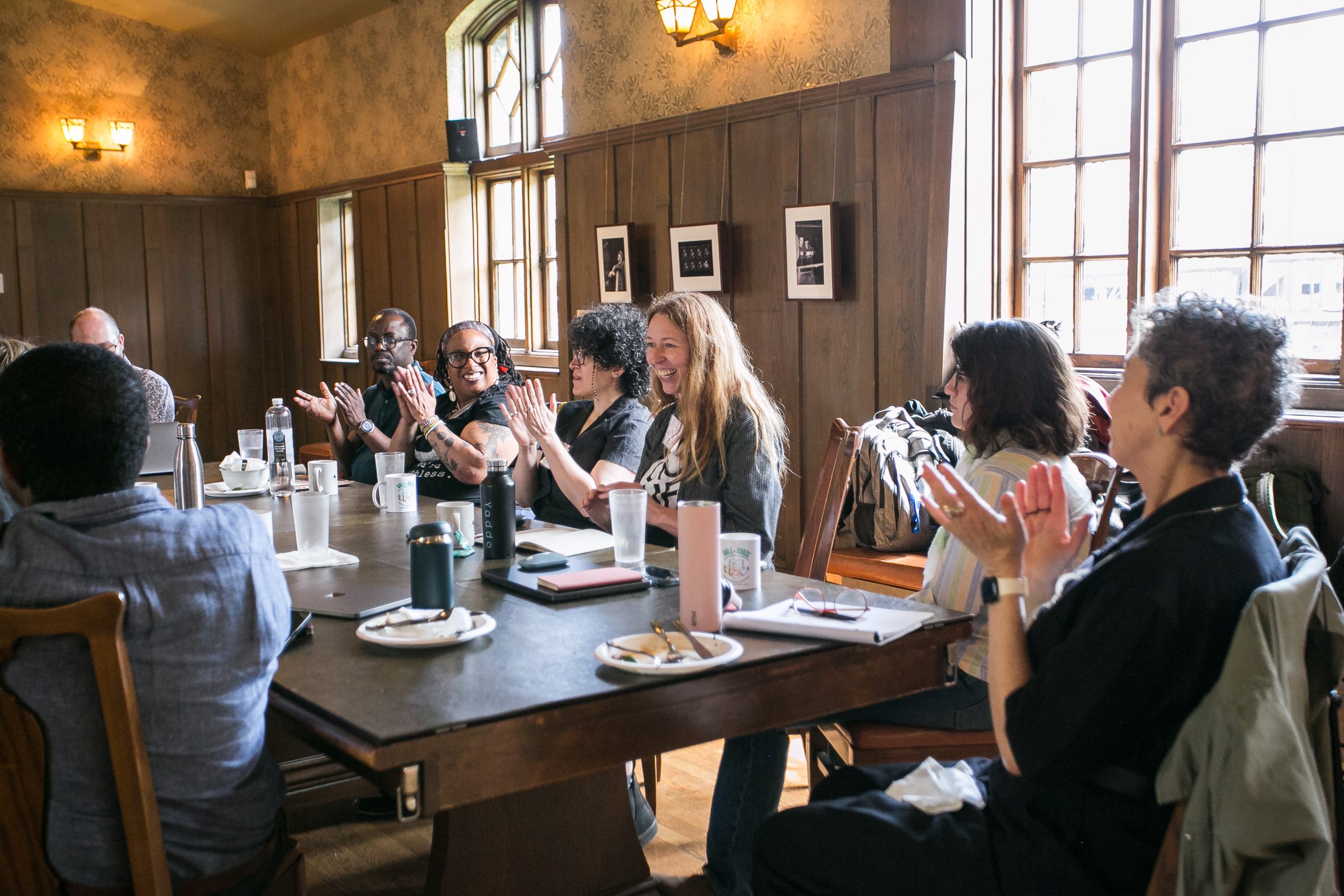 People sit around a table in a warmly lit room, smiling and clapping. Plates, cups, and notebooks are on the table, and sunlight streams in through large windows.