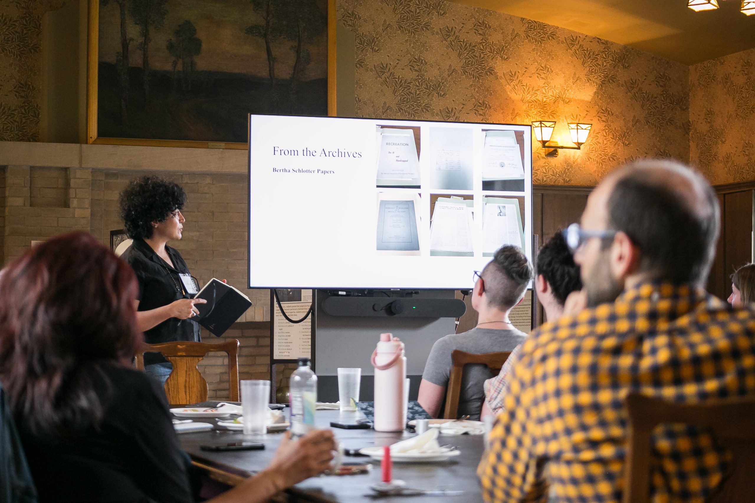 A woman presents archival documents displayed on a large screen to a group of people seated around a table in a warmly lit room.