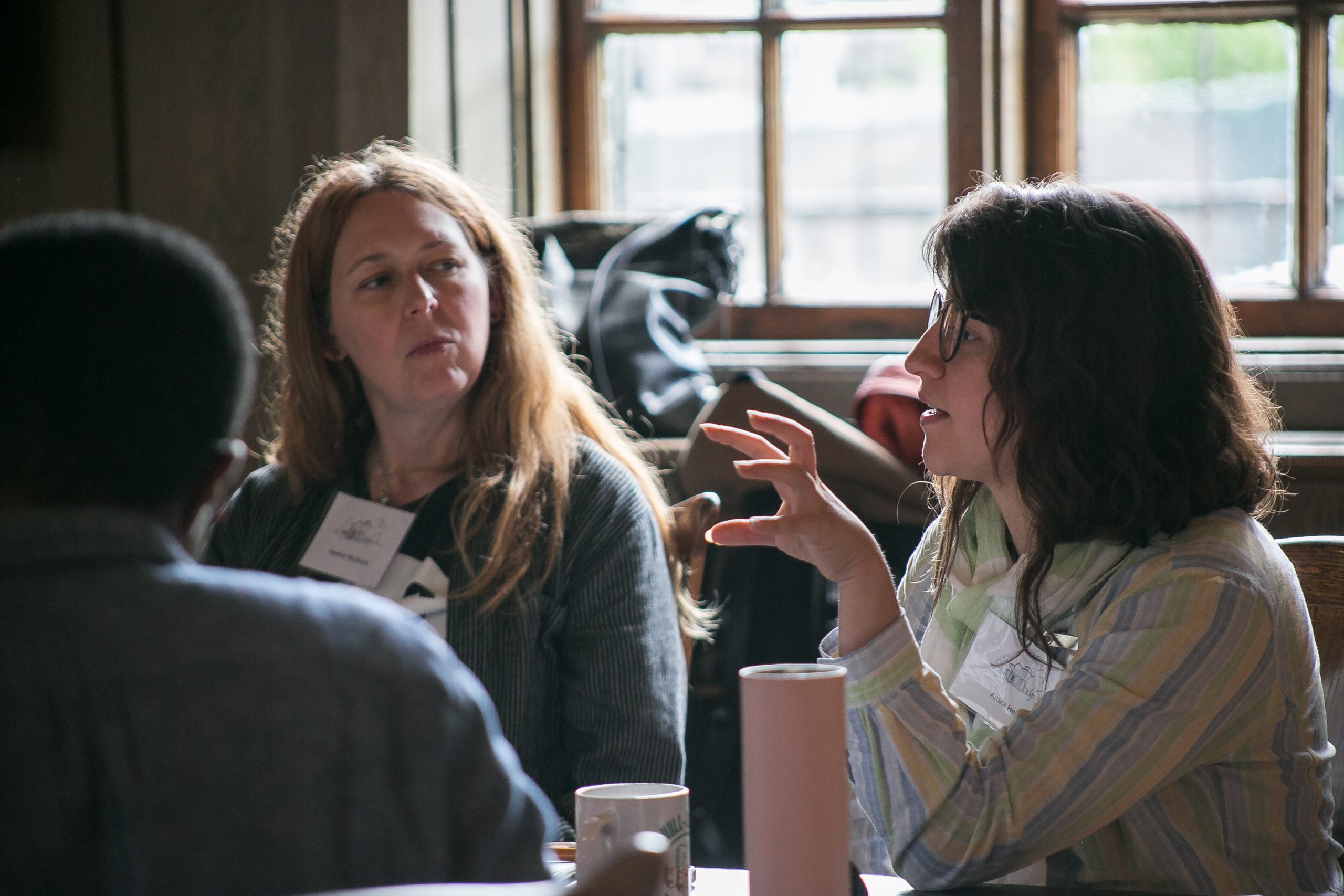 Three people sit at a table in conversation near a window. A woman with long hair listens while another gestures with her hand. Name tags and cups are visible. Light from the window illuminates the scene.