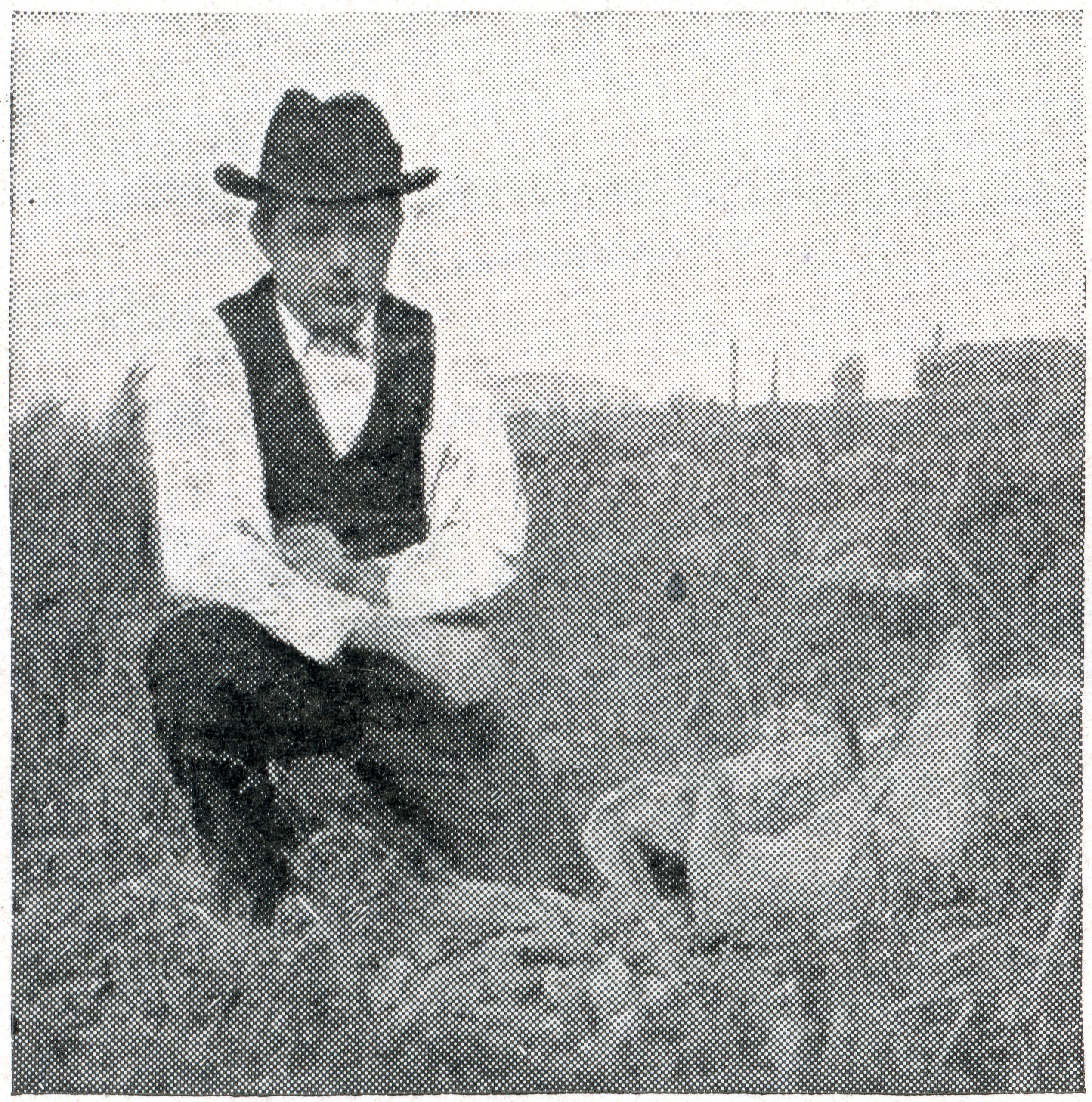 A man in a hat, vest, and bow tie kneels in a field with crops. Industrial buildings are faintly visible in the distant background. The image is black and white with a grainy texture.