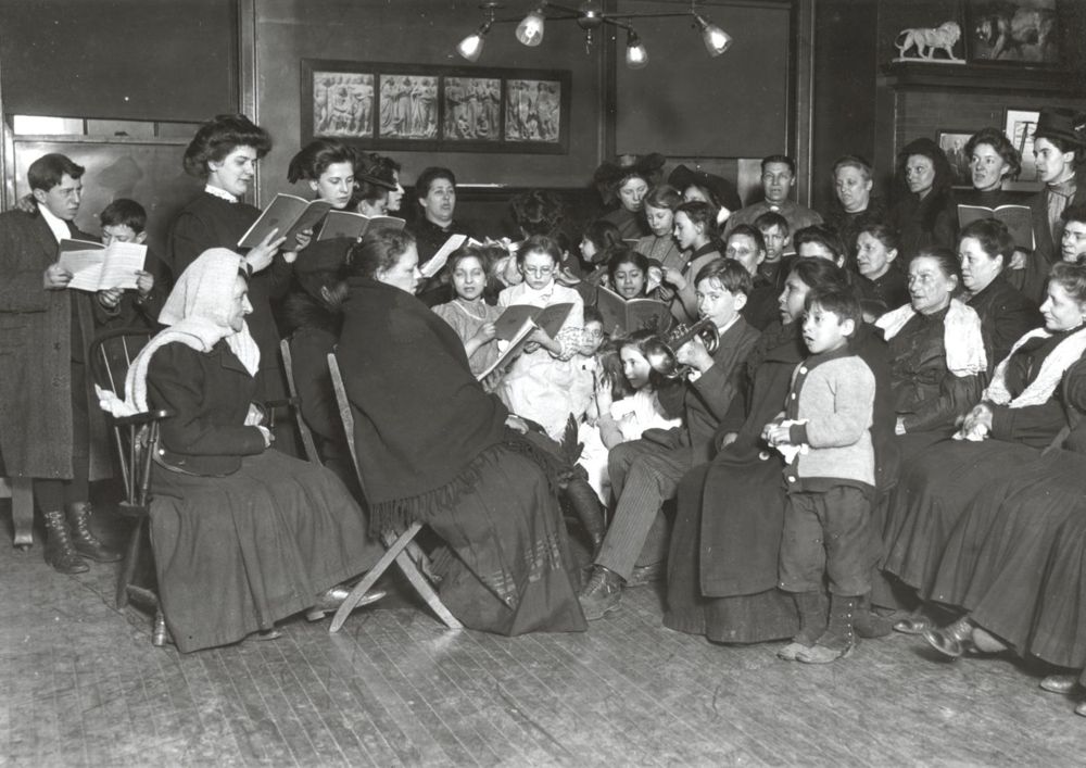 A group of men, women, and children sit and stand closely together in a crowded room, reading from books or papers, possibly singing or listening during a community gathering or class in an early 20th-century setting.