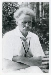 An older man with white hair and a mustache sits outdoors, wearing a light-colored shirt and scarf. He looks serious, with arms crossed, and leafy trees are visible in the background.