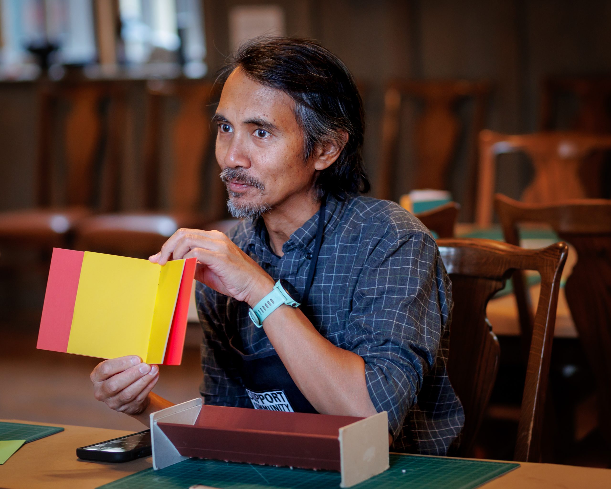 Artist Regin Igloria, a man with long hair and a beard holds a handmade book with red and yellow covers, sitting at a table with craft materials in a workshop setting.