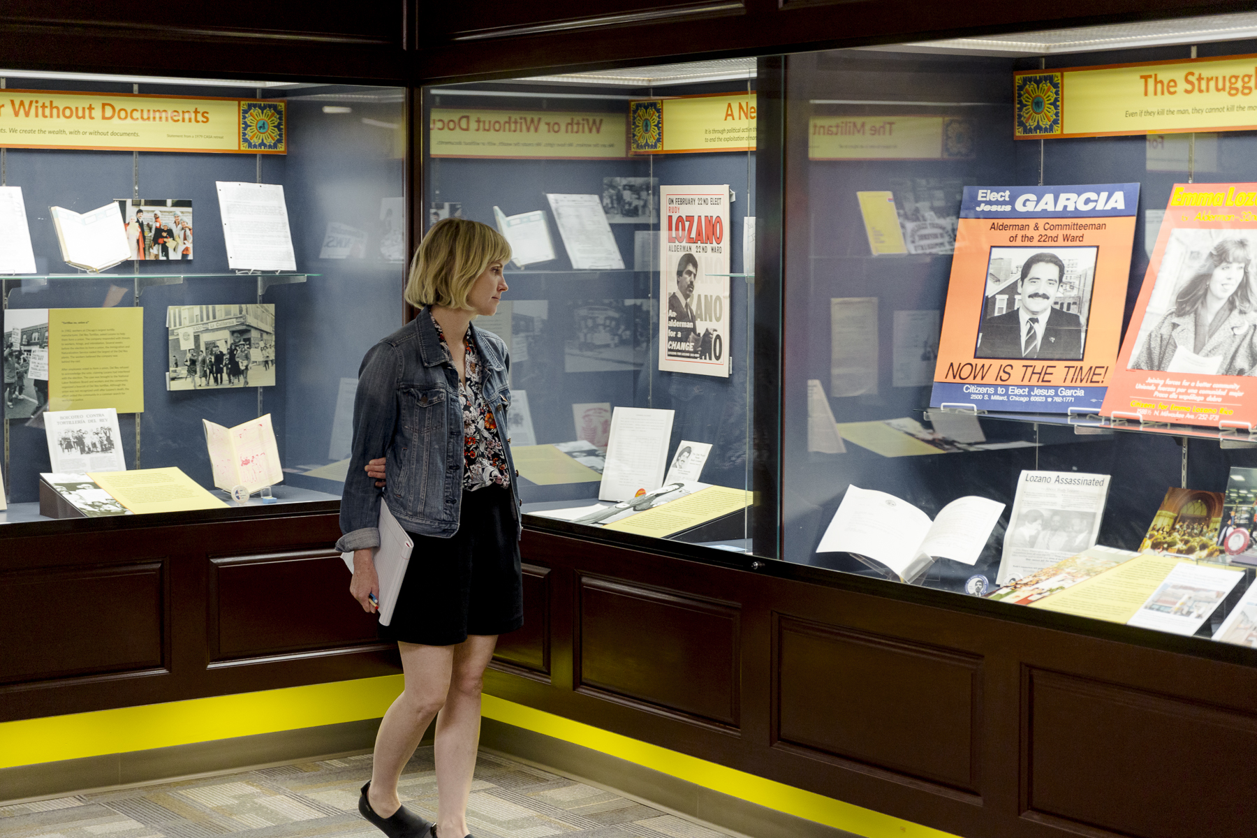 A woman views a museum display case filled with historical documents, photos, books, and posters, including a prominent campaign poster and materials about community activism.