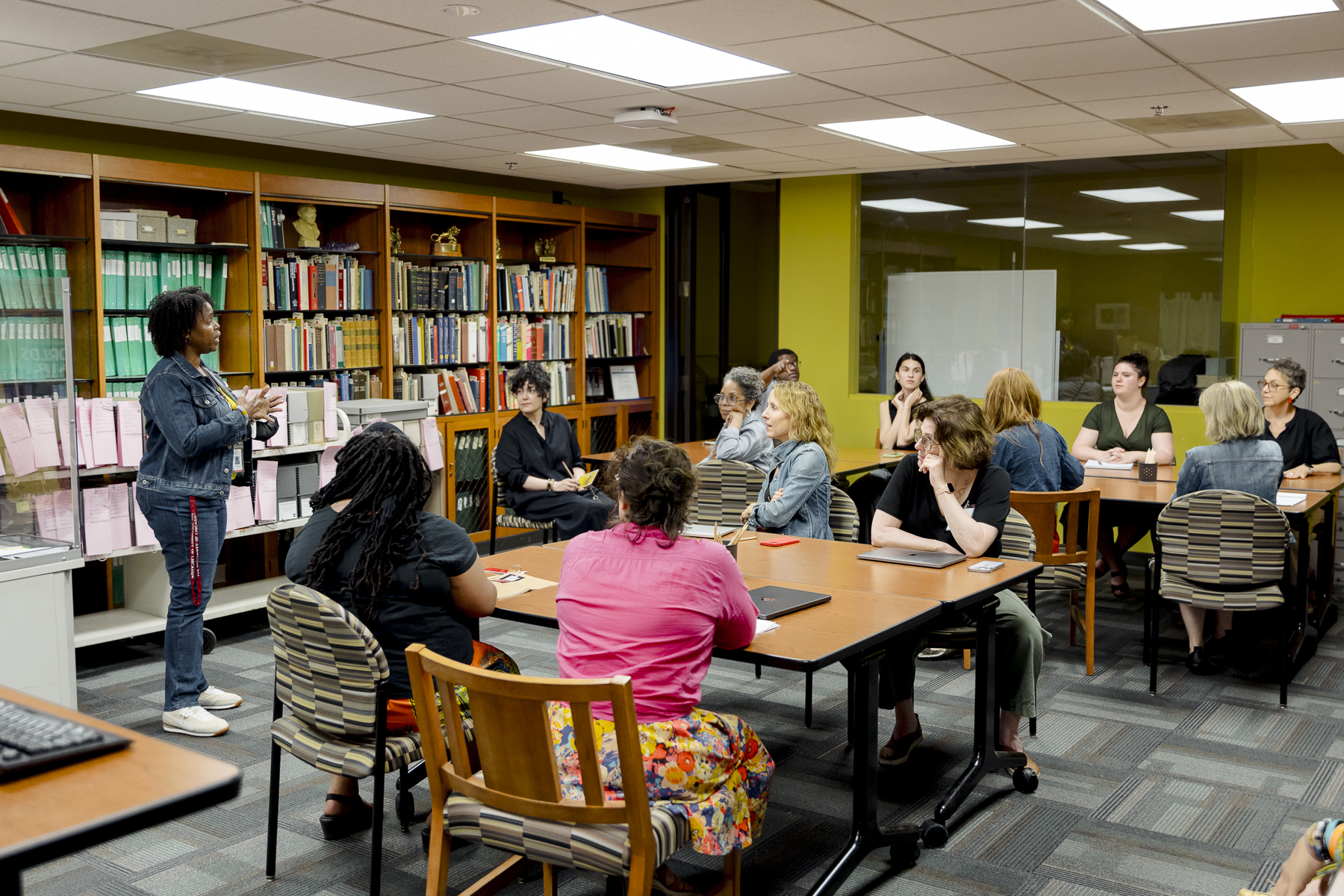 A woman stands and speaks to a group of adults seated at tables in a library or classroom, with bookshelves and files in the background.