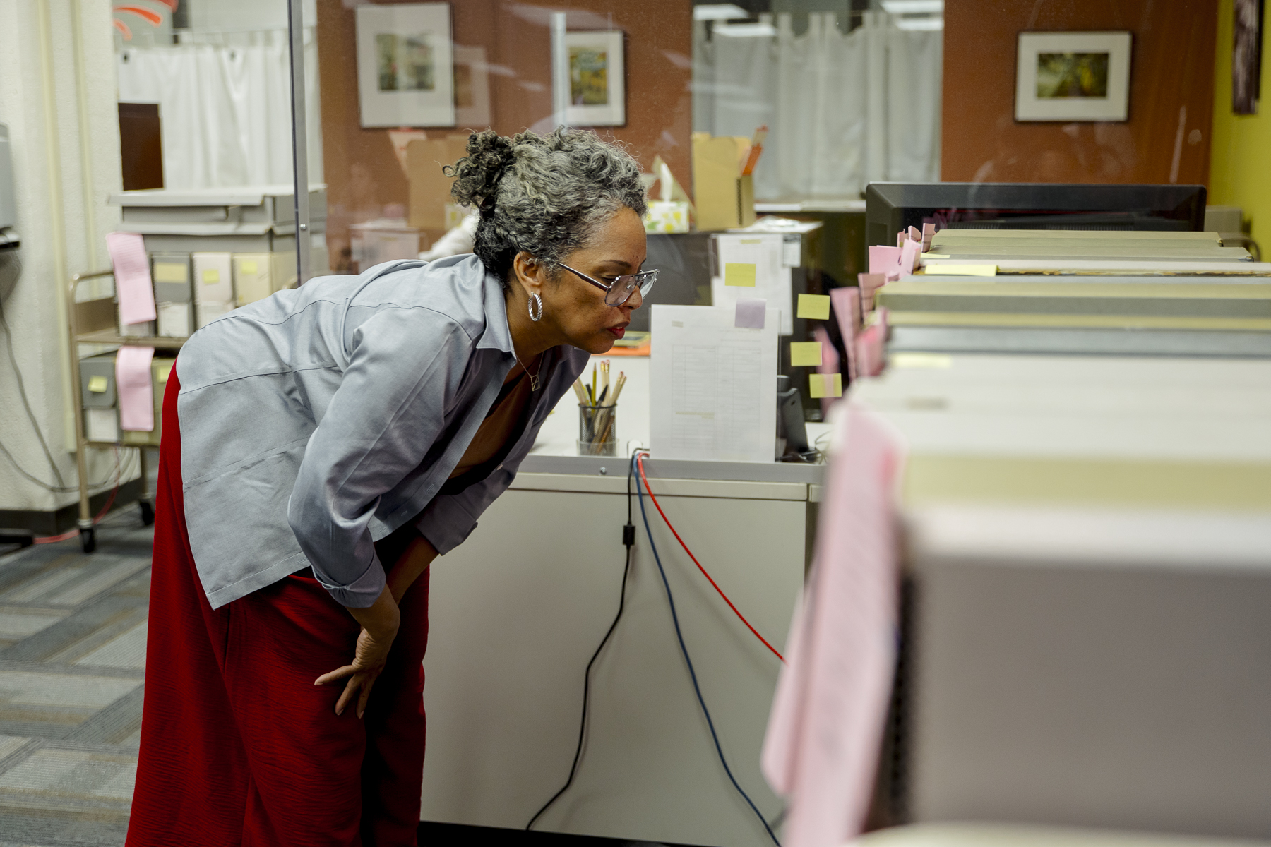 A woman with glasses and gray hair bends forward to look closely at a row of file folders in an office setting. Pink notes are attached to the files, and documents and photos are visible in the background.