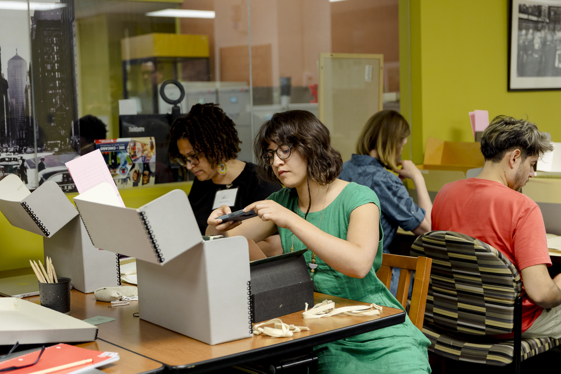 Several people sit at tables working with archival boxes and documents in a brightly-lit room with yellow walls and large windows. One woman in the foreground examines a black item closely.
