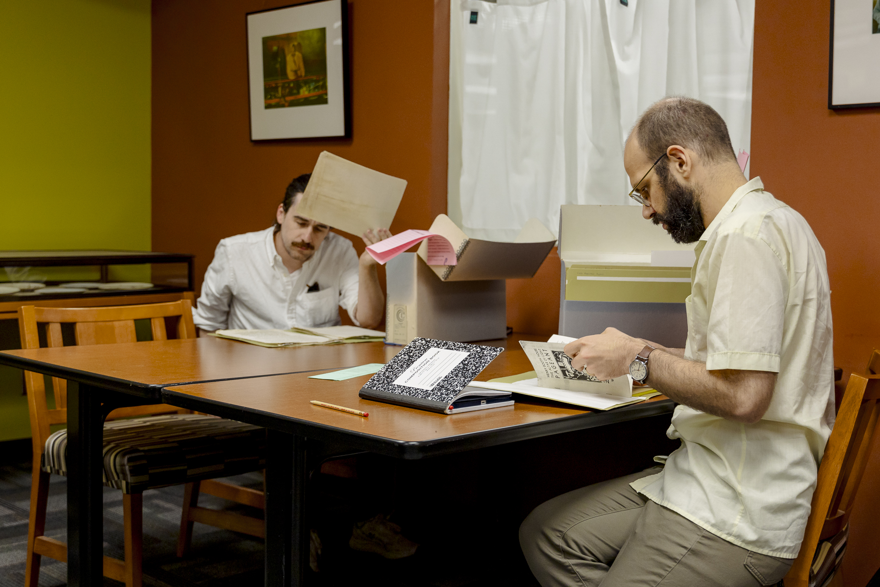 Two men sit at a table in a library or archive, examining documents and folders. One man writes in a notebook, while the other looks at papers from a box. A pencil and notebook are on the table.