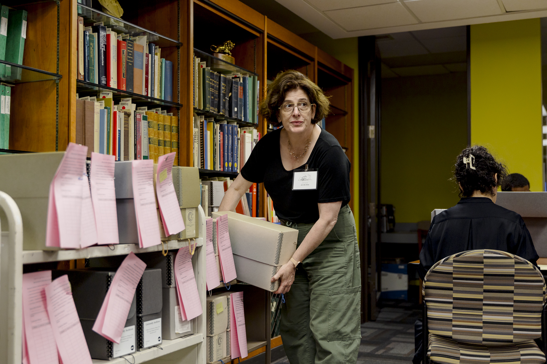 A woman with glasses stands by shelves filled with books and archive boxes, holding a box. Pink slips are attached to many boxes. Another person sits at a table nearby, working.