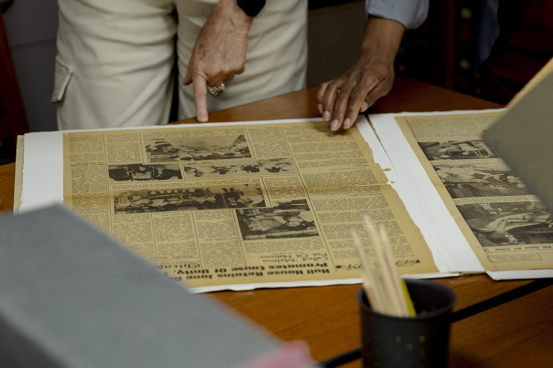 Two people examine vintage newspapers laid out on a table. One person points at an article while the other rests a hand on the paper. The newspapers feature black and white photos and text.