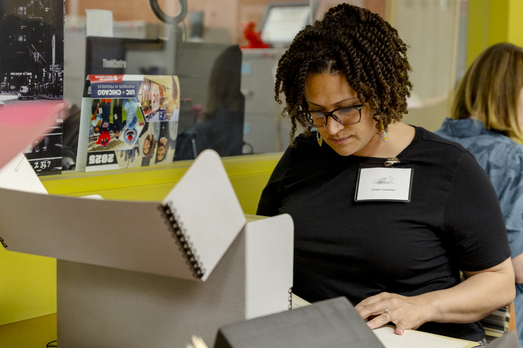 A woman with curly hair and glasses reads documents from an archival box at a table. She wears a black shirt and a name tag. Another person works at a nearby desk. The background includes a yellow wall and various items.