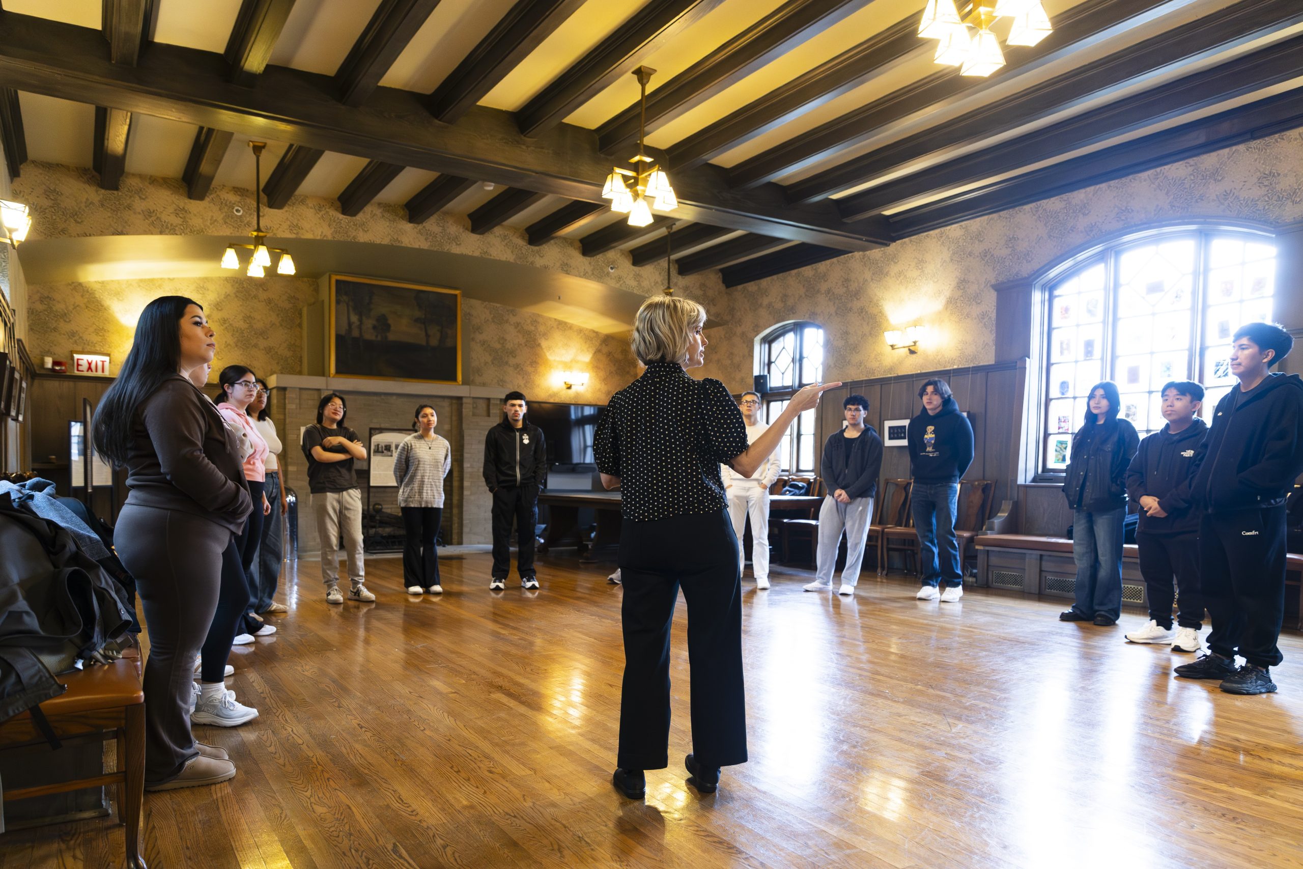A woman stands in the center of a wooden-floored room, speaking to a group of people arranged in a circle. The room has large windows, wooden beams, and warm lighting.