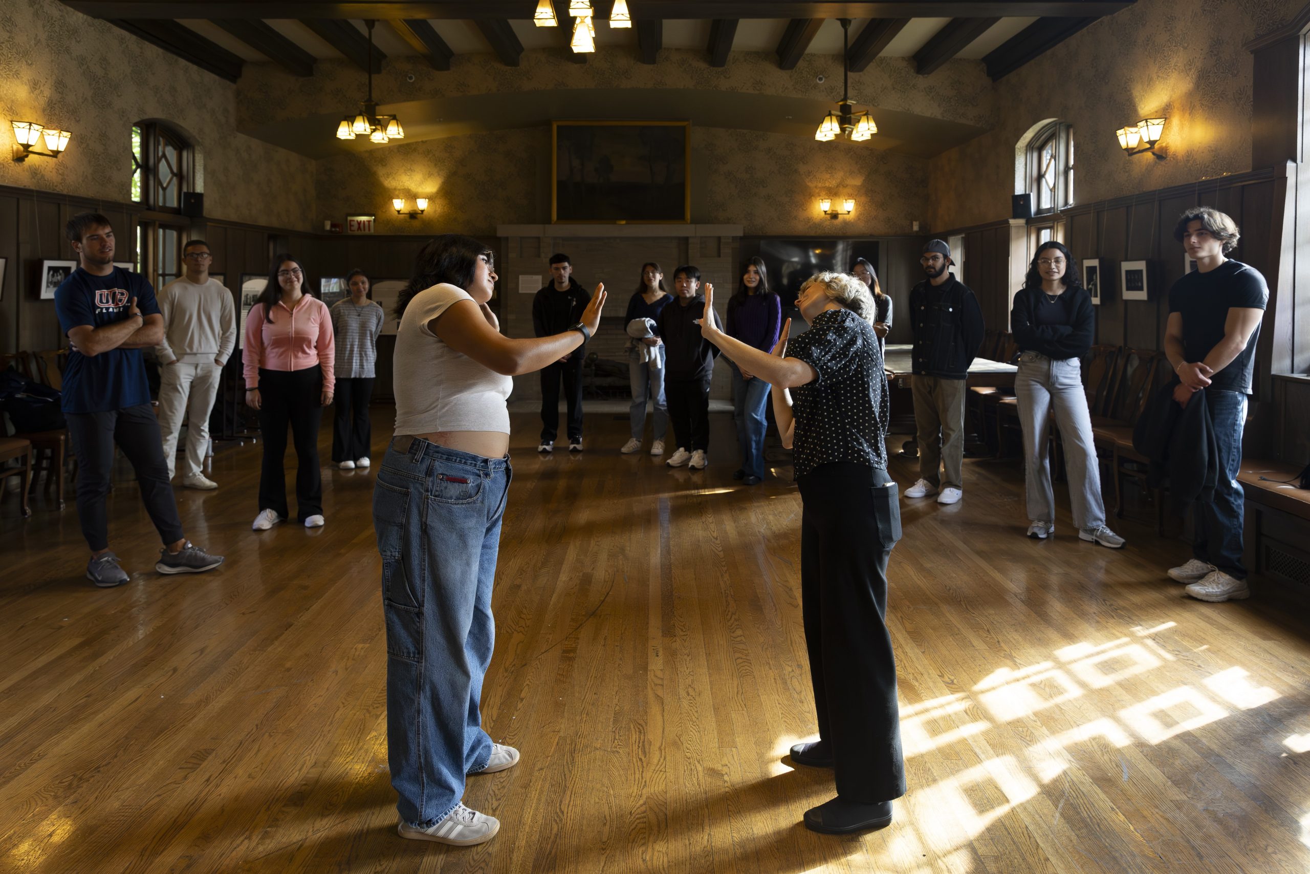A group of people stands in a circle in a wood-paneled room while two individuals in the center face each other with raised hands, appearing to participate in an interactive activity.