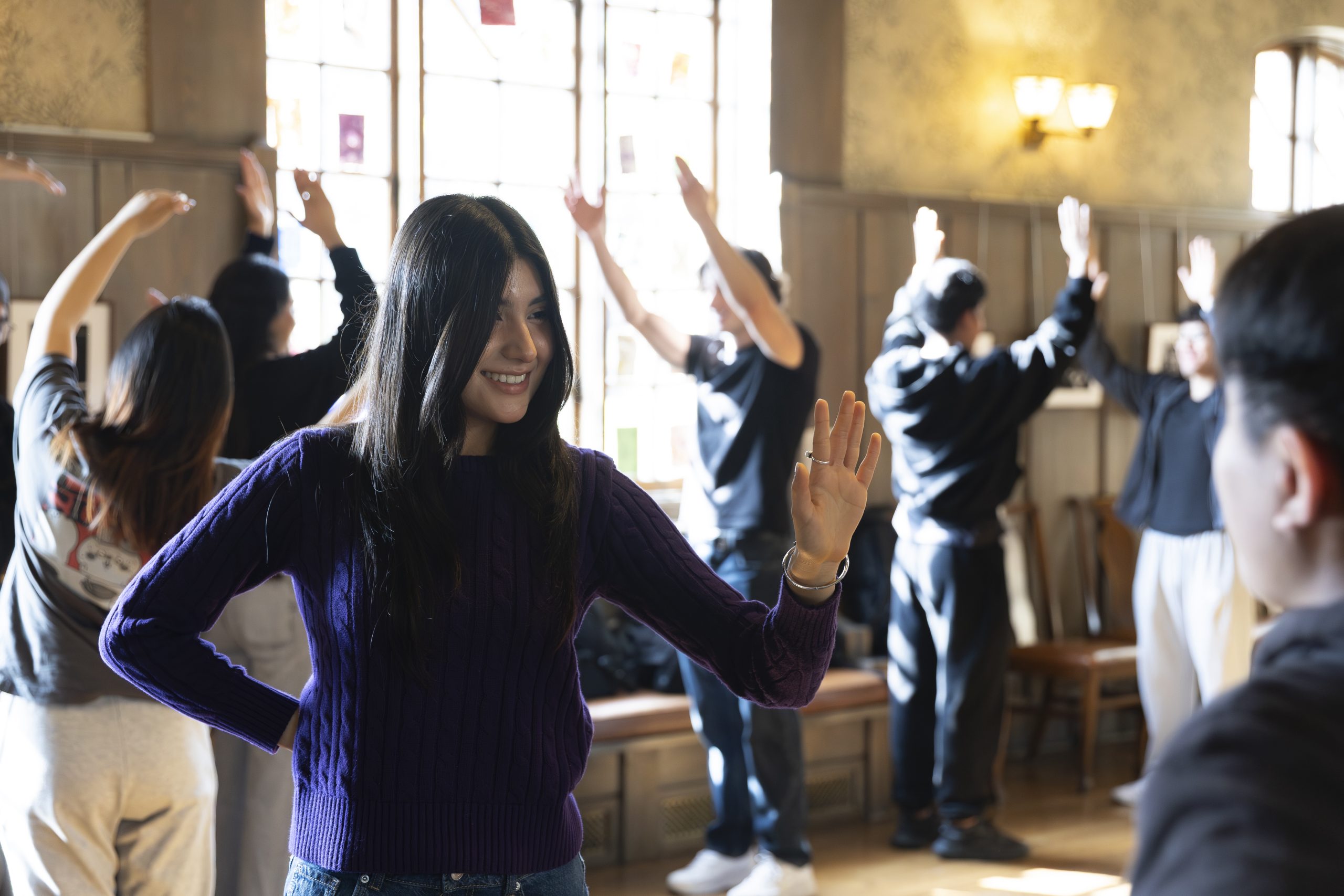 A woman in a purple sweater smiles and raises her hand, engaging with another person. Several people in the background stand in pairs, also raising their hands, in a sunlit room.