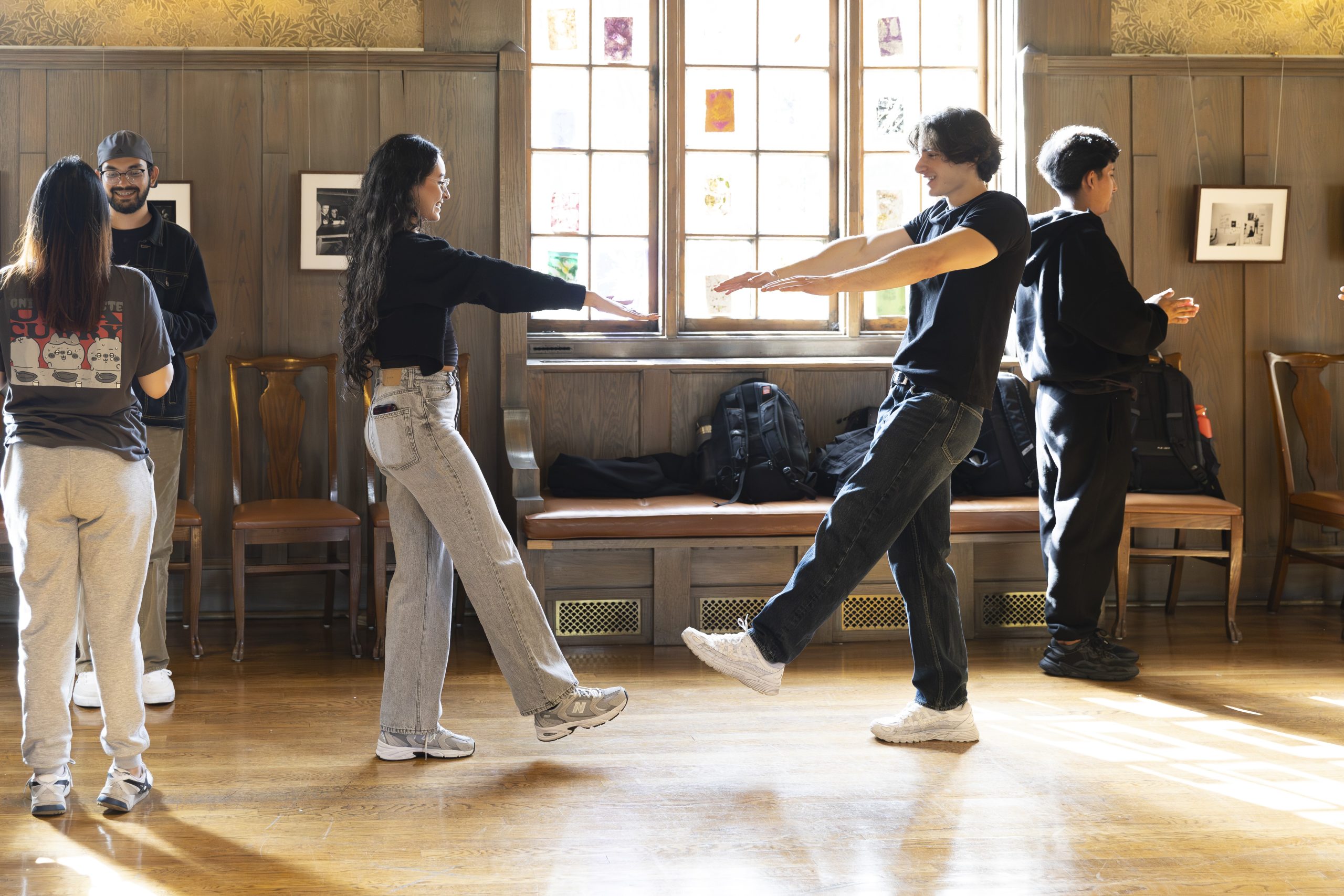 Six people stand in pairs in a sunlit room with wooden walls, talking and laughing. Two pairs face each other with arms outstretched and one leg raised, while another pair stands near the window chatting.