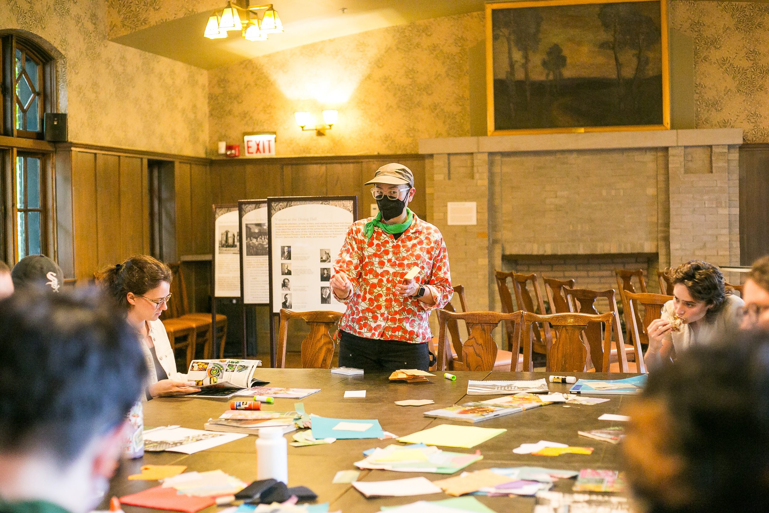 Artist, Olly Costello. A person wearing a mask and floral shirt stands and speaks to a group seated around a table covered with colorful papers in a wood-paneled room.