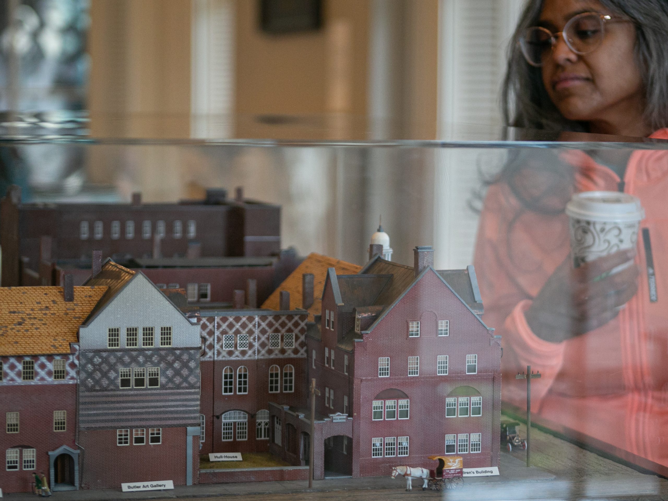 A person holding a coffee cup looks at a detailed miniature model of old brick buildings displayed inside a glass case.