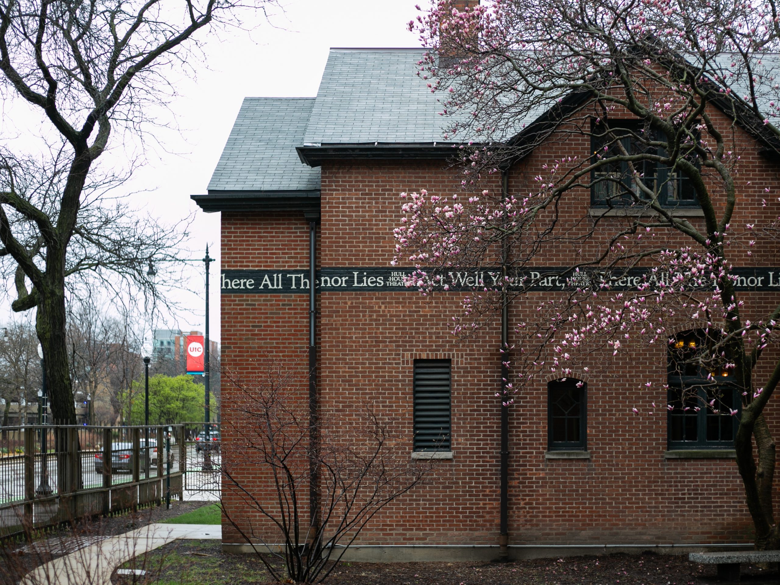 A brick building with blooming pink flowers on a tree in front, bare trees nearby, and a partially visible quote running along the side of the building. The scene appears to be overcast and early spring.