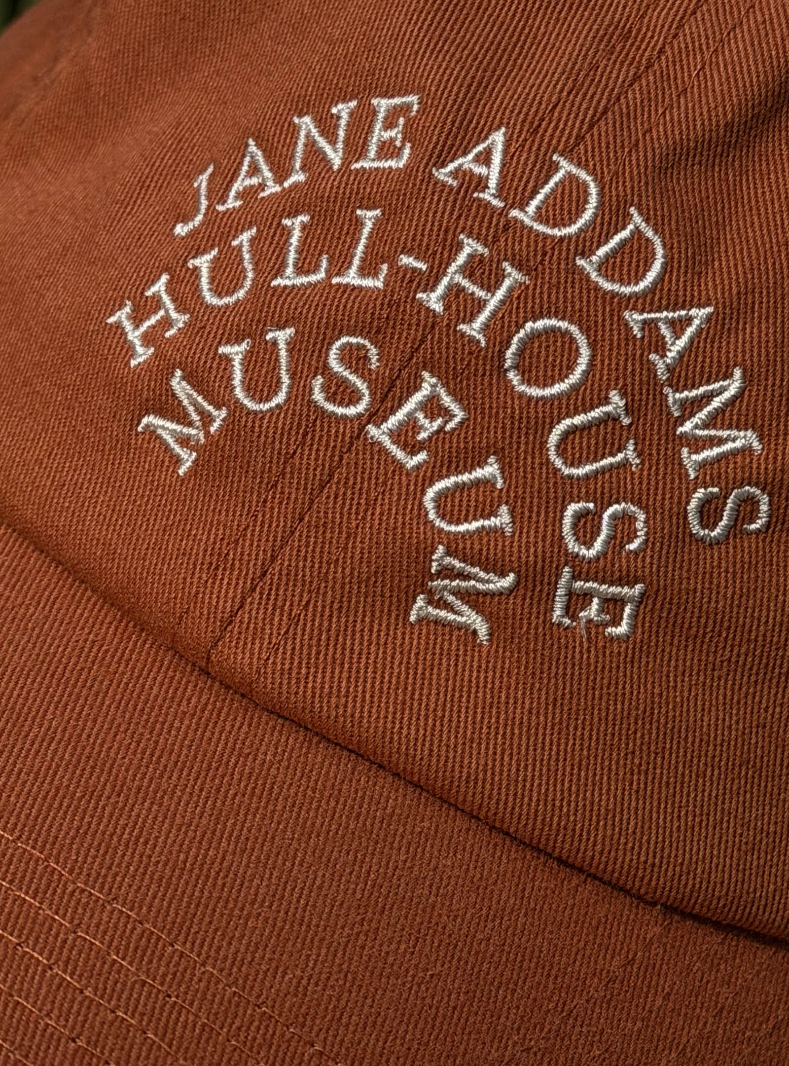 Close-up of a rust-colored fabric cap with Jane Addams Hull-House Museum embroidered in white, arched text.