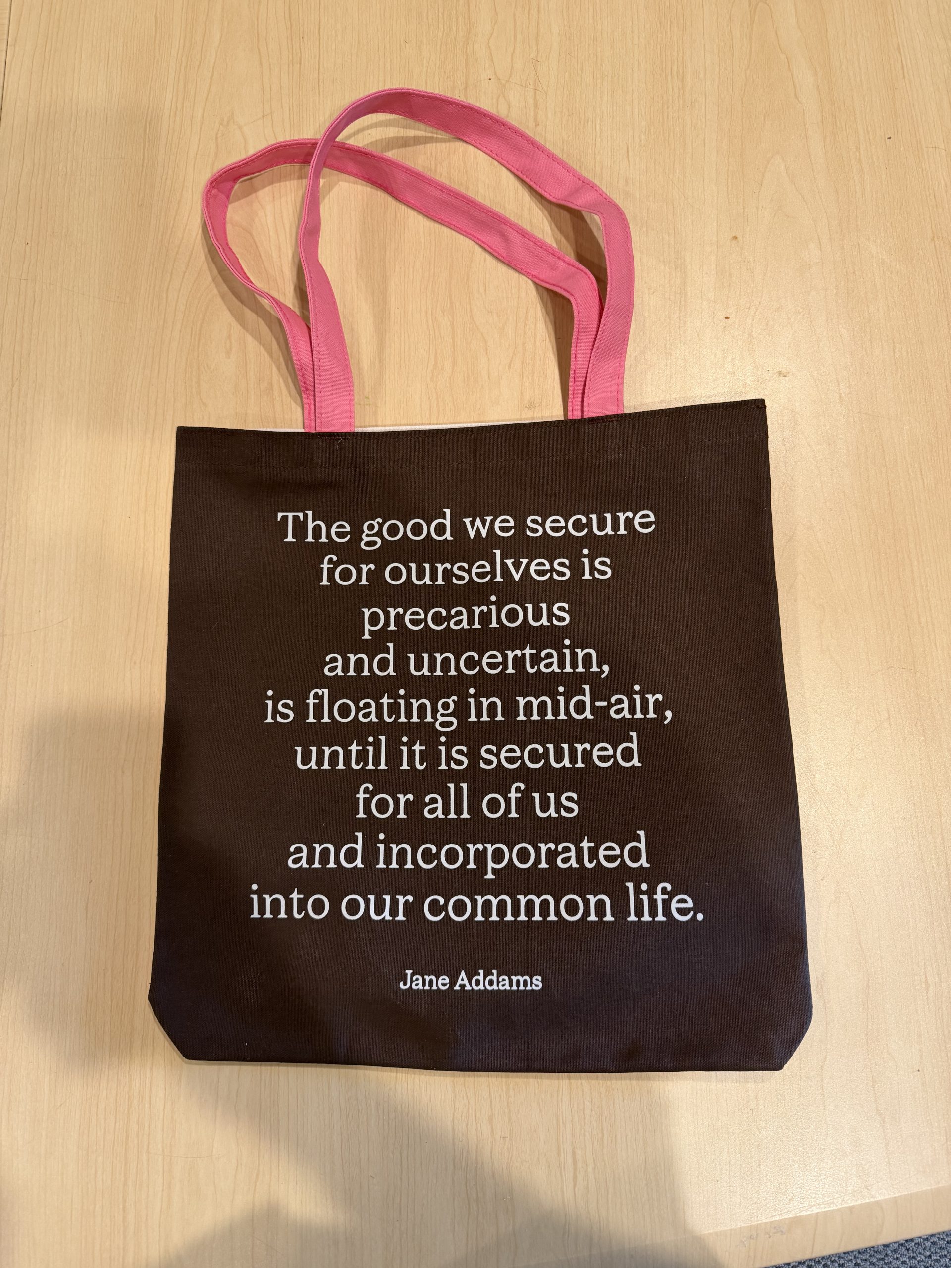 A brown tote bag with pink handles on a wood surface. The bag has a Jane Addams quote printed in white letters about securing good for ourselves and others for a common life.