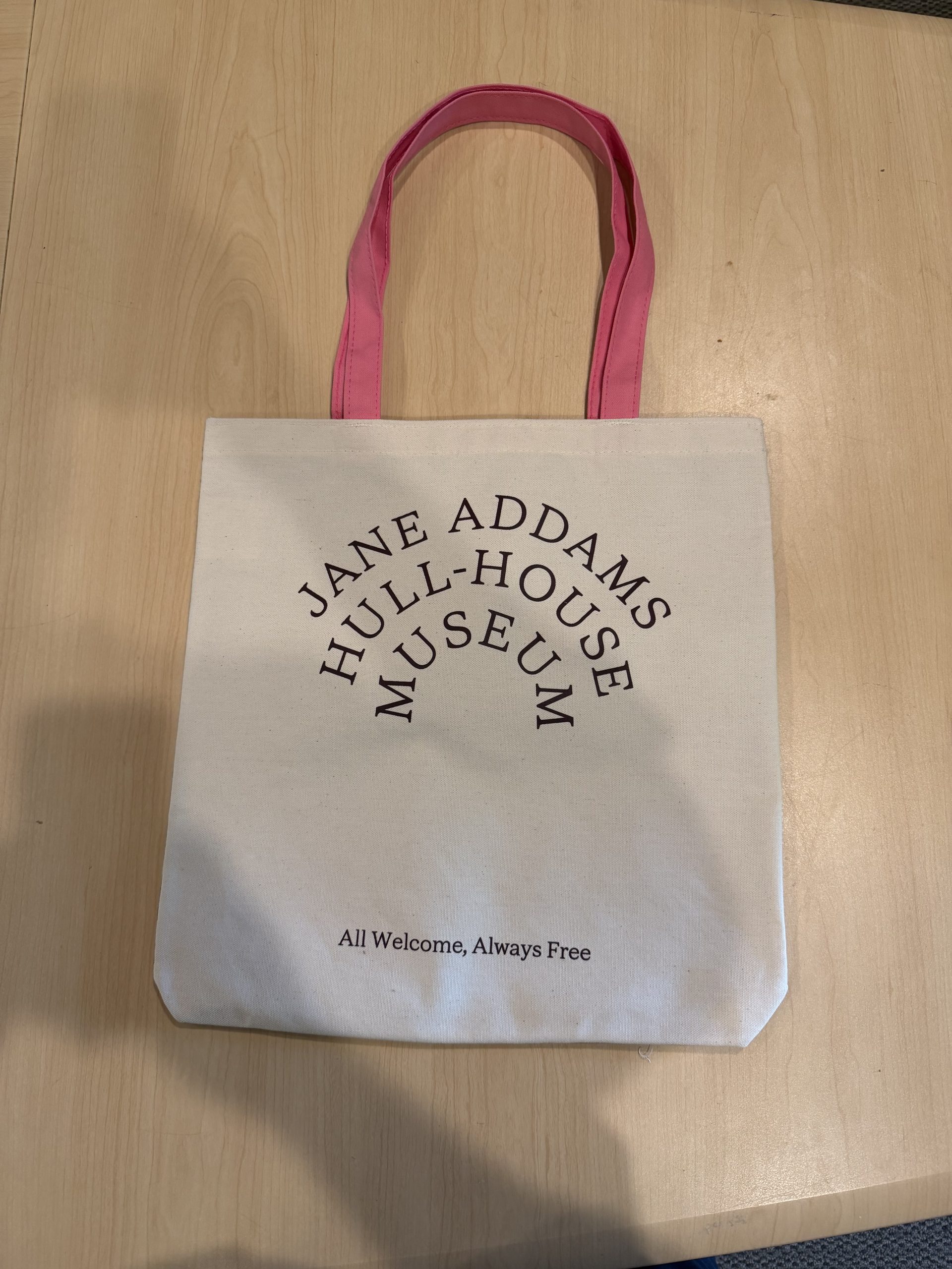 A white tote bag with pink handles reads Jane Addams Hull-House Museum in an arc and All Welcome, Always Free below. The bag is placed on a light wood surface.