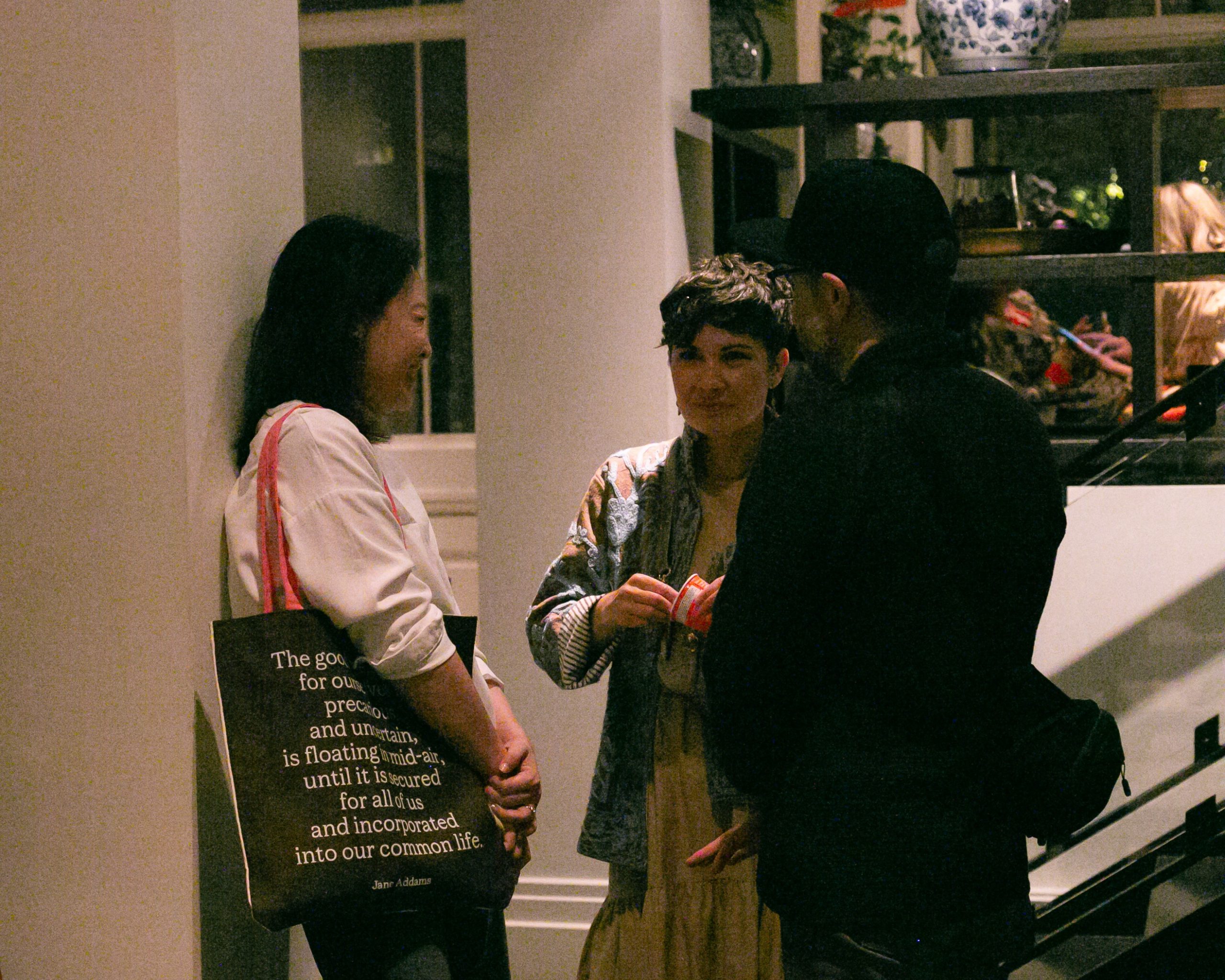 Three people stand in an indoor entryway, with a woman on the left carrying a black tote bag printed with a Jane Addams quote.